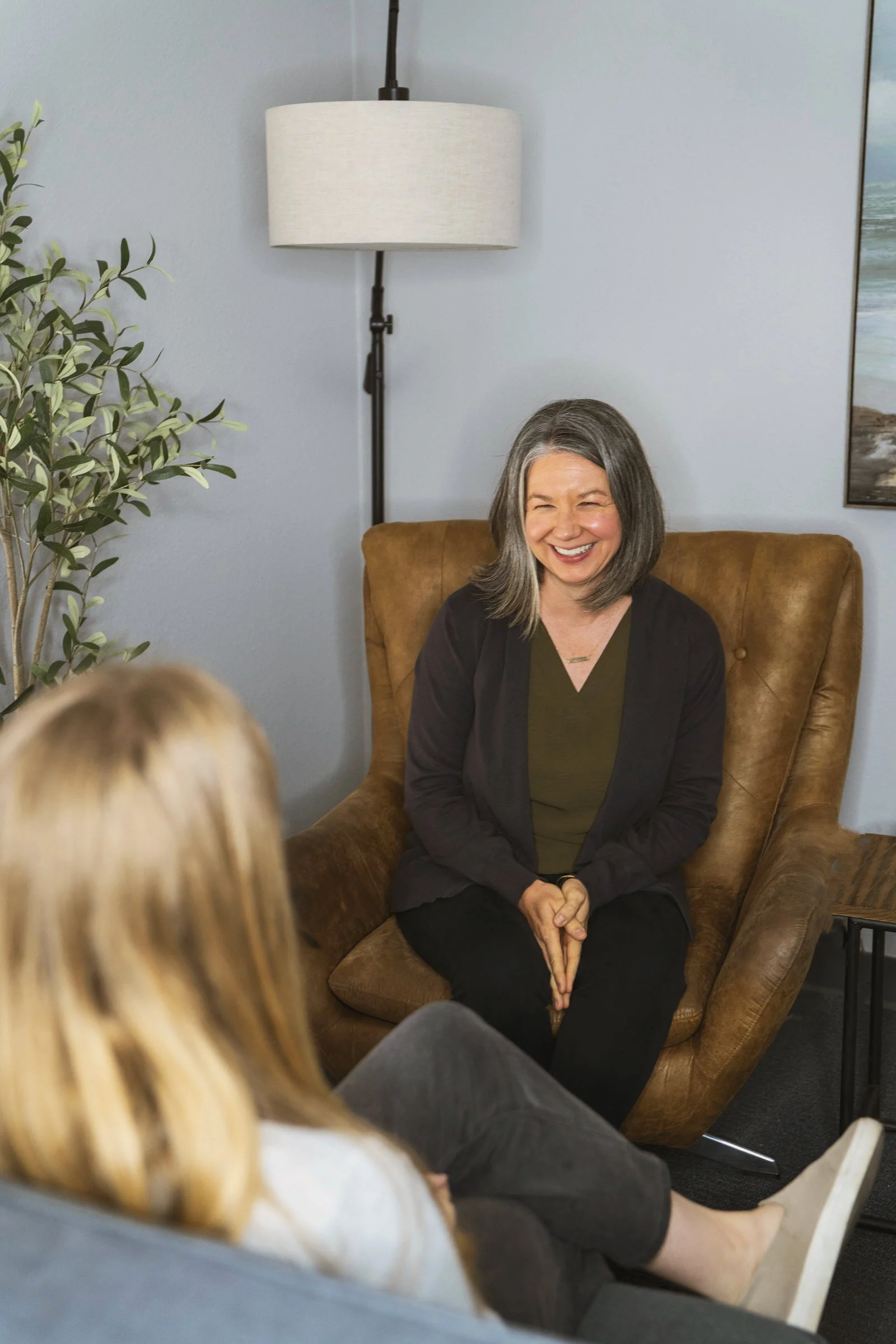 Two women having a conversation in an office, one sitting comfortably in a leather armchair and the other sitting on a couch with blond hair. The woman in the armchair is smiling and appears engaged.