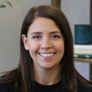 A woman with long brown hair smiling indoors, with a bookshelf and decorative item in the background.