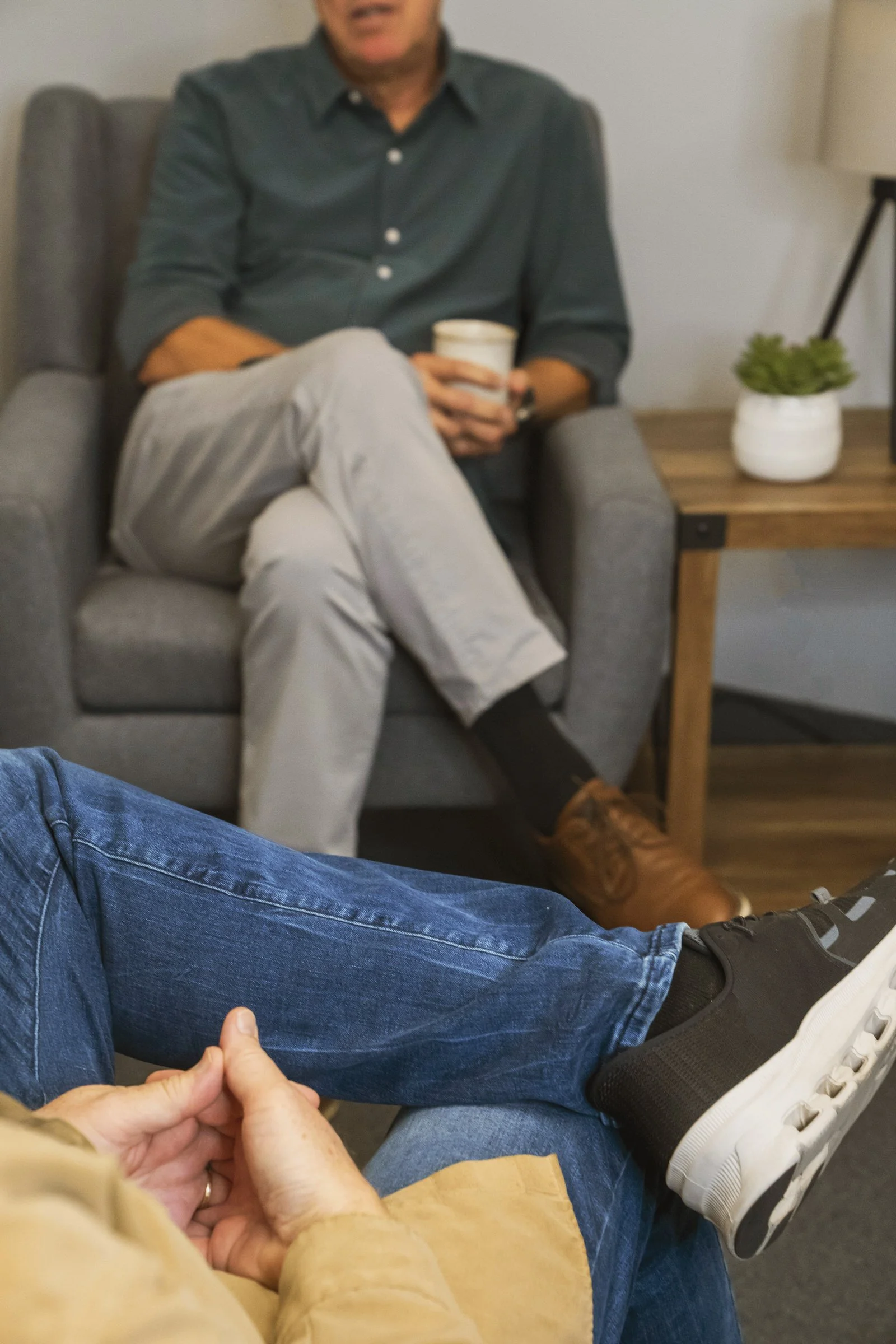 Two people are sitting and engaging in conversation. One person in the background is sitting on a grey armchair, holding a cup, dressed in a dark grey shirt and beige pants. The other person in the foreground is sitting with crossed legs, wearing blue jeans, a tan jacket, and black sneakers with white soles. There is a wooden side table with a small potted plant next to the armchair.