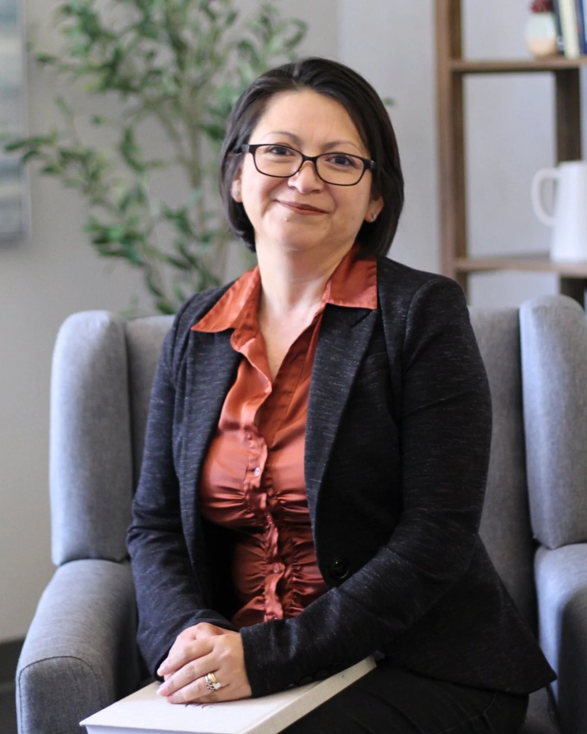 A woman with glasses and dark hair, wearing a black blazer and a rust-colored blouse, sitting in a gray armchair in an office setting.