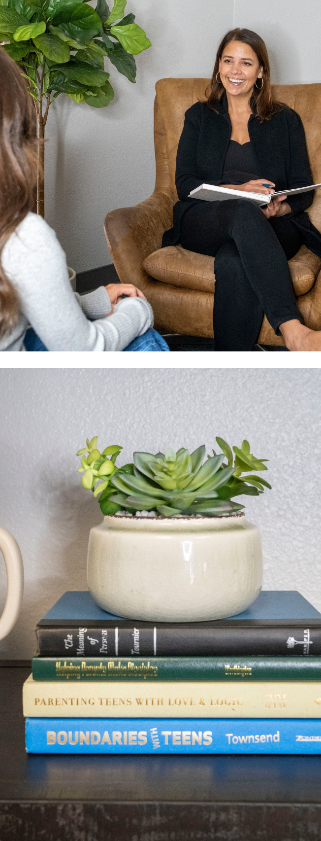 A woman sitting on a brown leather chair, smiling and holding a notebook, engaged in conversation with another woman whose arm and part of her head are visible. There is a green houseplant behind her and a table below displaying a stack of books and a succulent plant in a beige pot.