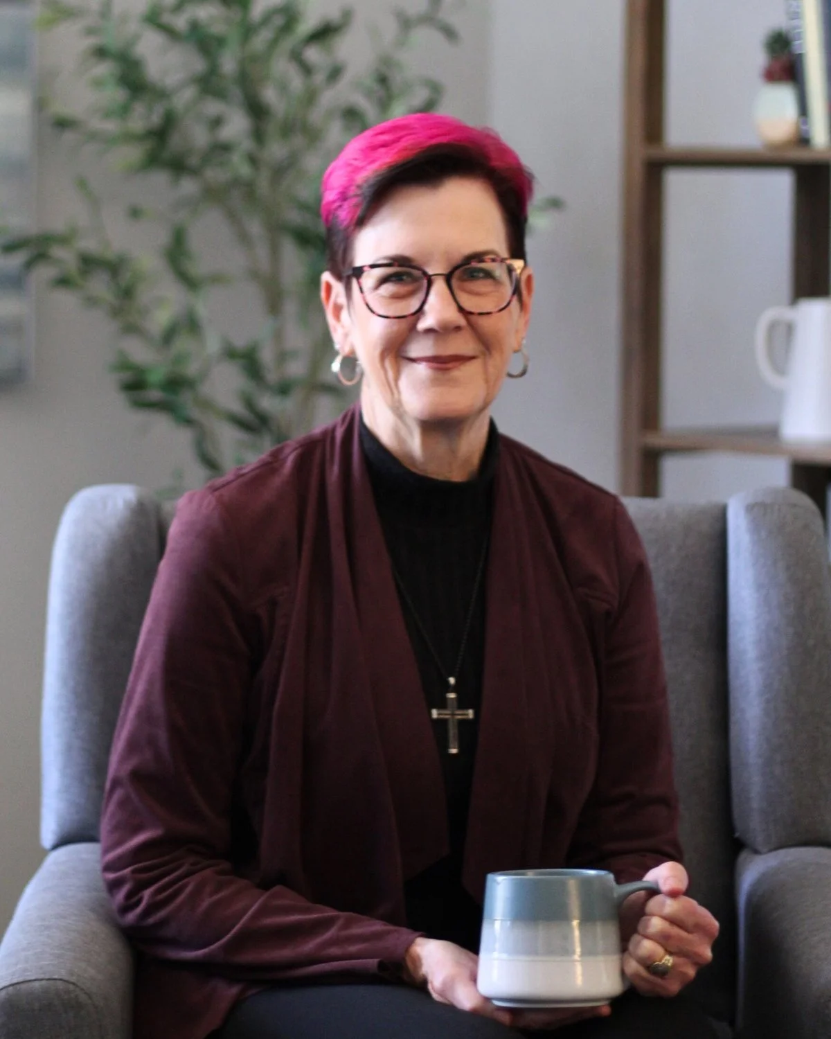 A woman with short hair dyed pink at the tips, wearing glasses, a black turtleneck, and a maroon blazer, sitting on a gray chair indoors, holding a mug, smiling.