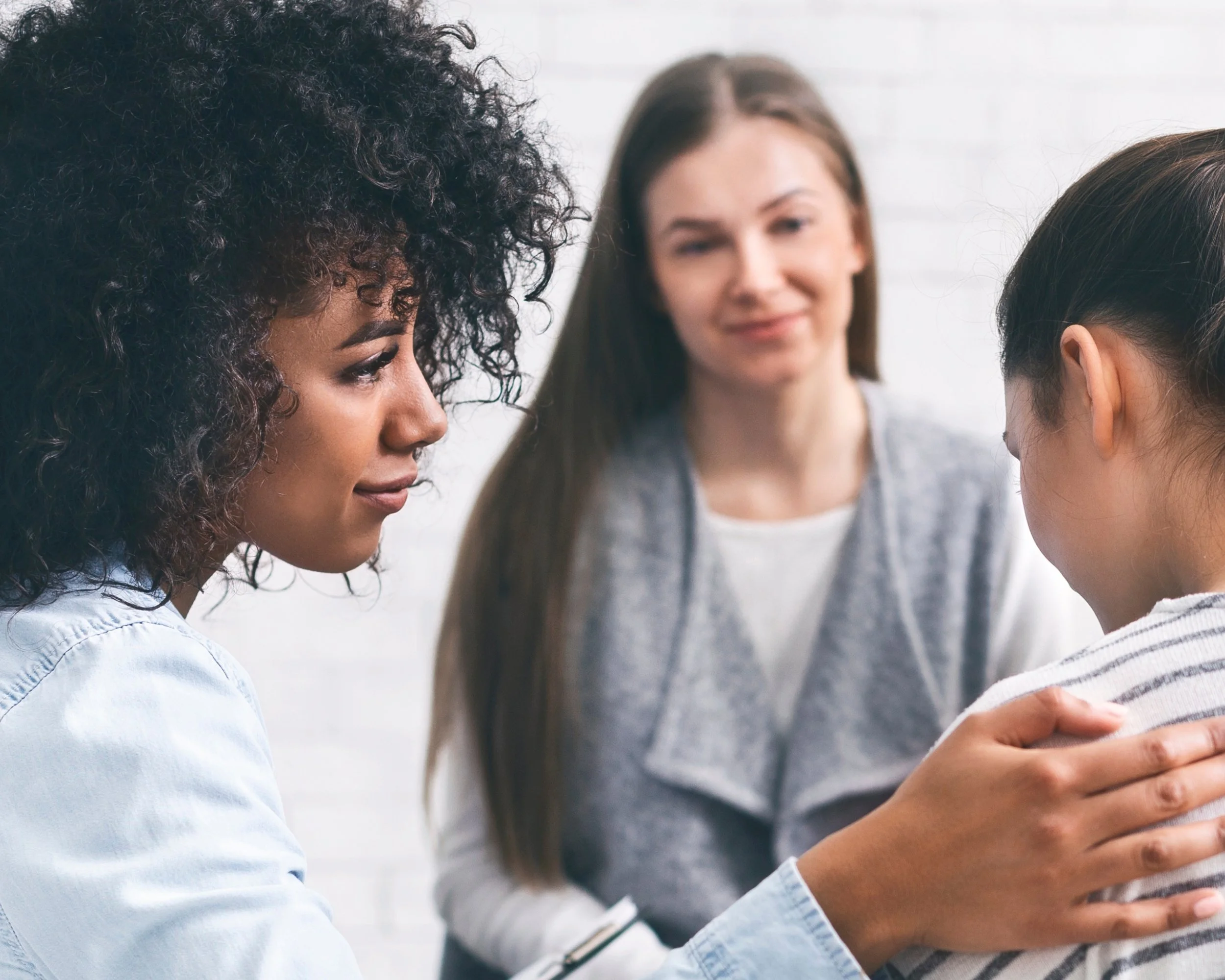 Three women in a supportive interaction, with one woman gently holding another woman's shoulder in a clinical or counseling setting.