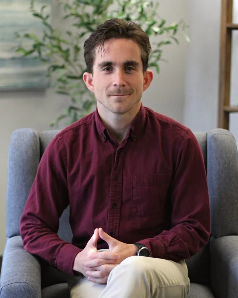 A man with brown hair, dressed in a maroon button-up shirt and light-colored pants, sitting on a gray armchair with hands clasped, in a room with a plant and bookshelf in the background.