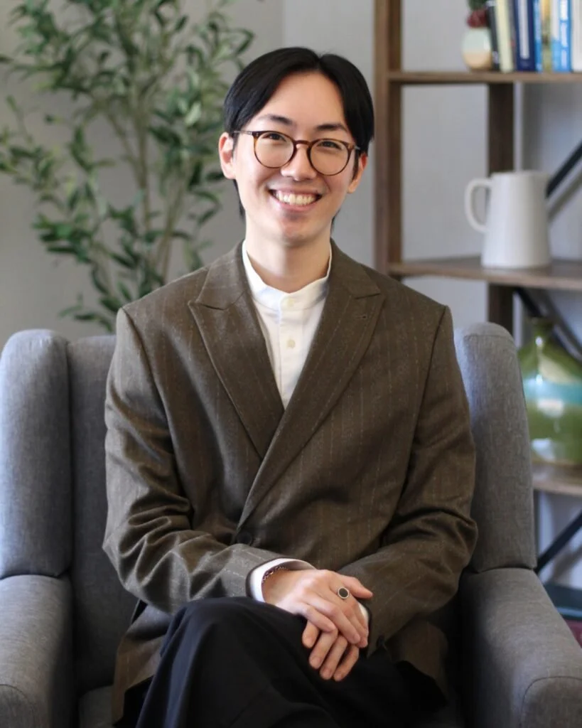 A smiling young man with black hair and glasses, wearing a brown blazer and white shirt, sitting on a gray couch in a cozy, modern office with bookshelf and green plant in the background.