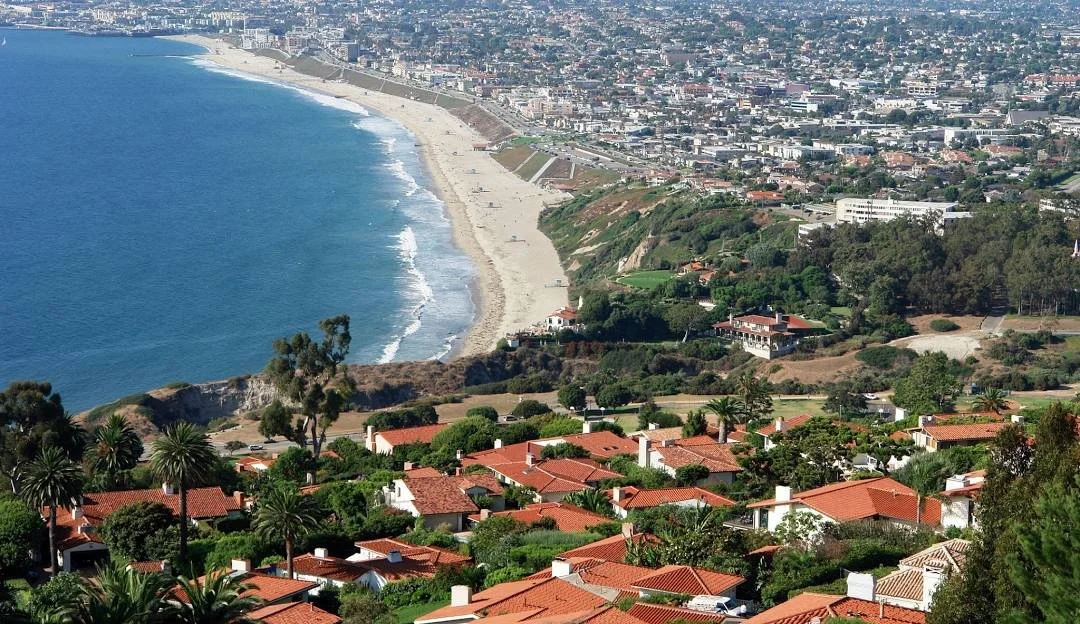 Aerial view of a coastal city showing a beach, ocean, and residential area with red-tiled roofs, trees, and green landscape.