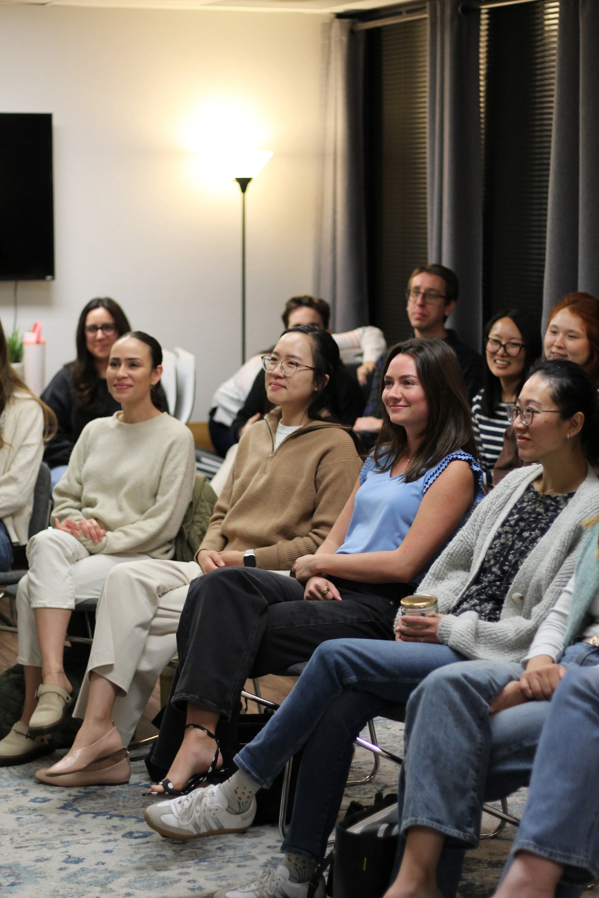 A group of diverse people sitting and smiling during a presentation or event in a room with gray curtains, a floor lamp, and a wall-mounted TV.