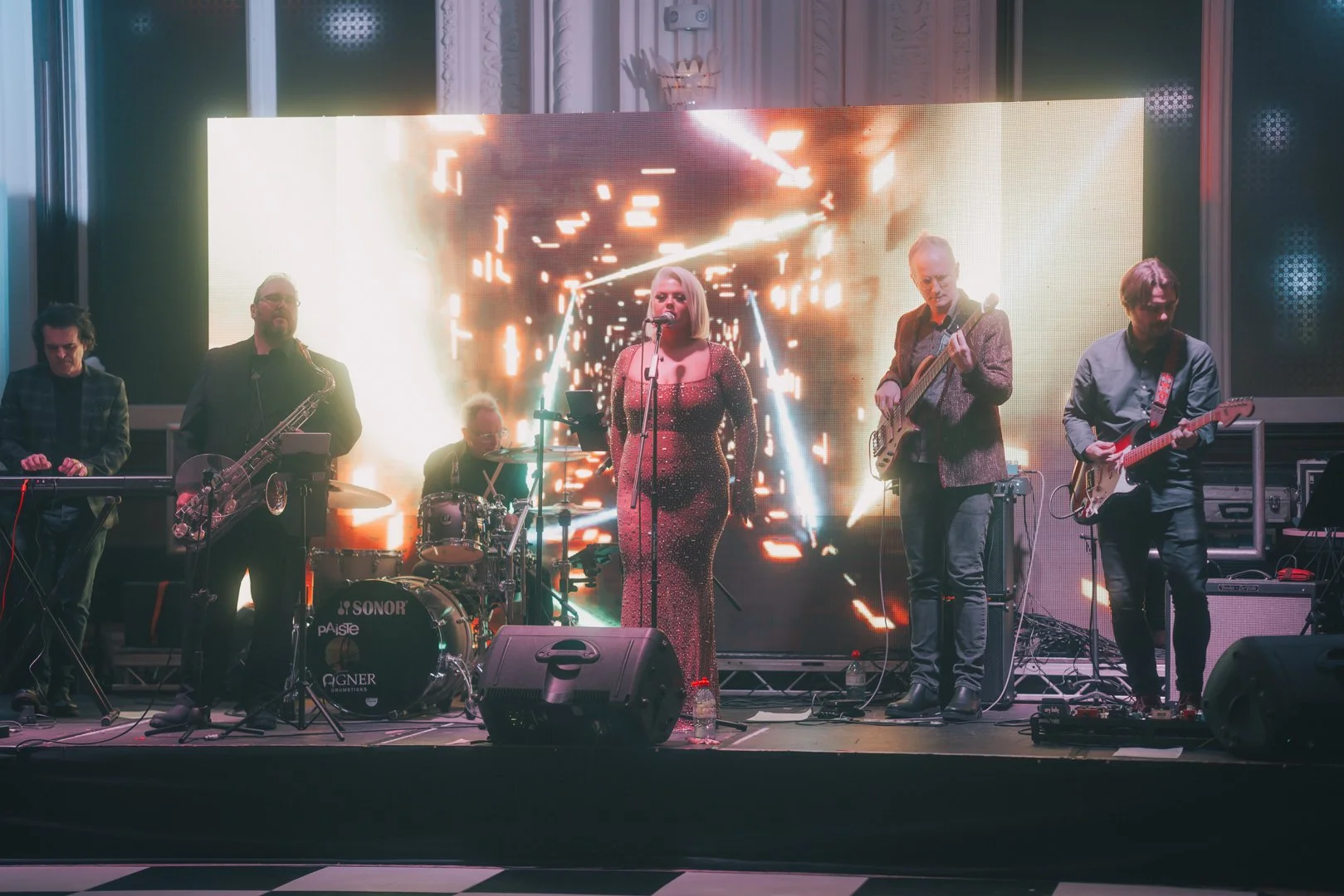 A band performing live on stage, featuring a female lead singer in a sparkling dress, a drummer, a keyboardist, and two guitarists, with a large digital screen displaying abstract visuals in the background.