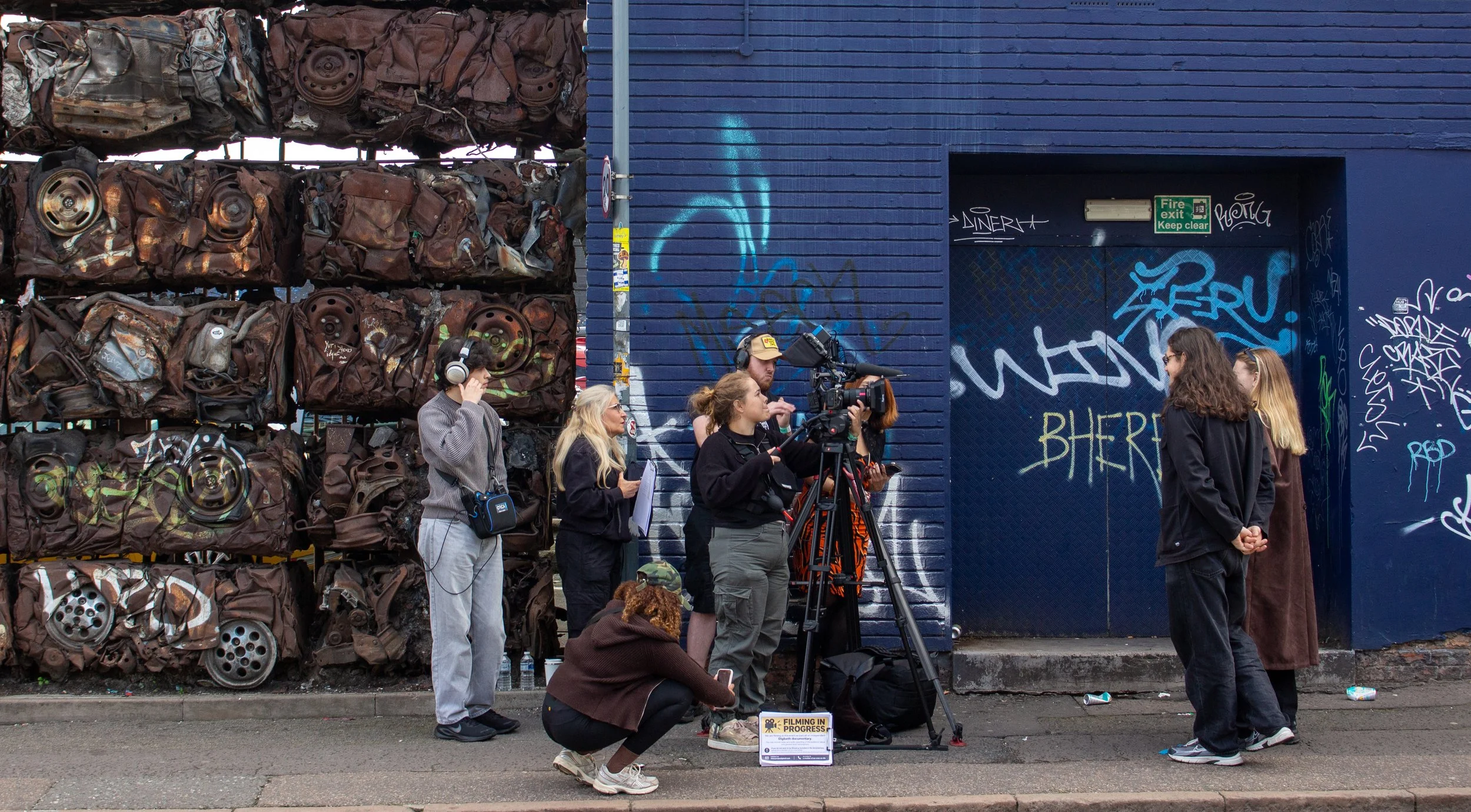 A group of people filming and taking photos on a city street, with a large wall of scrapped car parts on the left and a blue brick building with graffiti on the right.