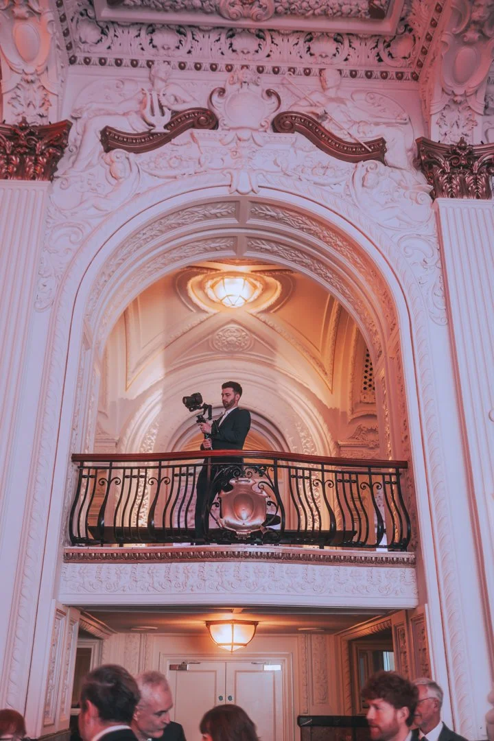 A man filming with a professional camera on a balcony of an ornate, elegant room with intricate architectural details.