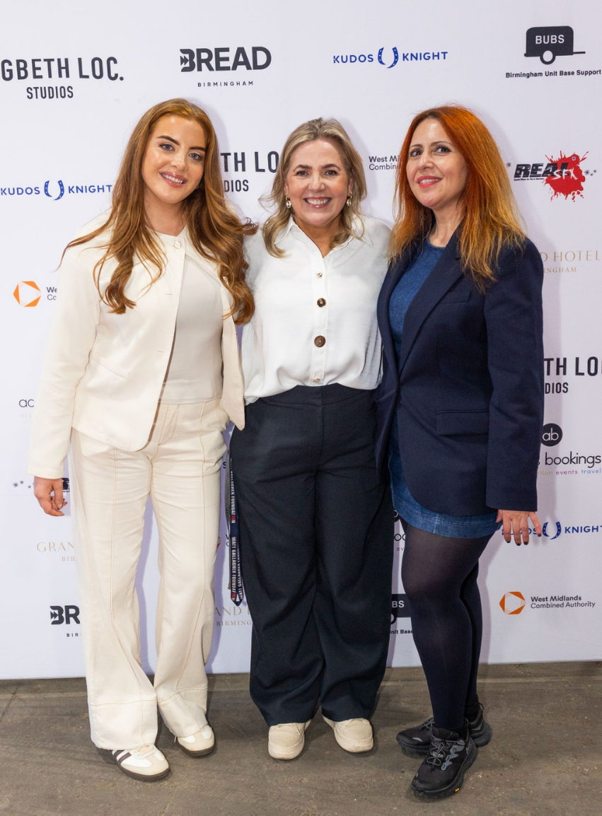 Three women standing together at an event, smiling. The backdrop behind them features various sponsor logos.