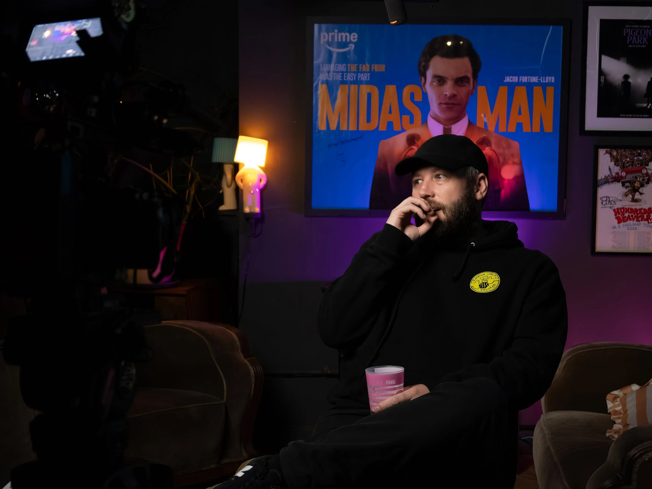 A man with a beard wearing a black cap and hoodie is sitting in a dimly lit room, holding a pink cup, with a large poster of the movie 'Midnight Man' featuring a man with dark hair and a pink shirt in the background.