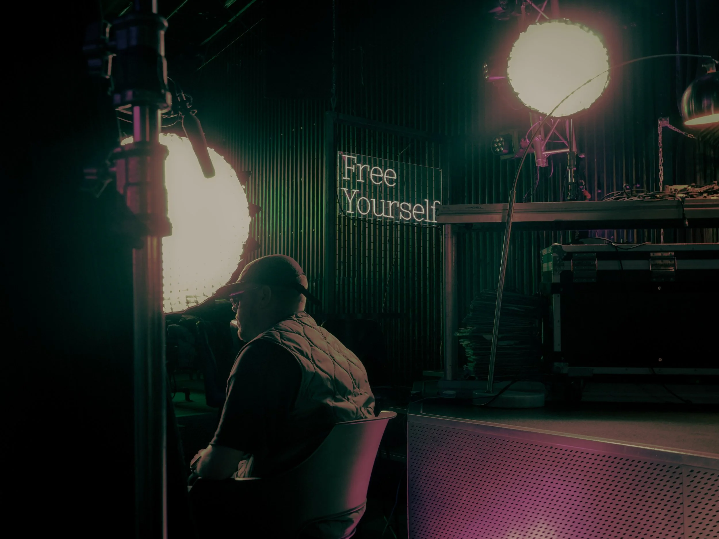 A person with glasses and a jacket sitting in a chair, facing away. The background has neon lights, a lit sign reading 'Free Yourself,' and stage lighting equipment illuminating the scene.