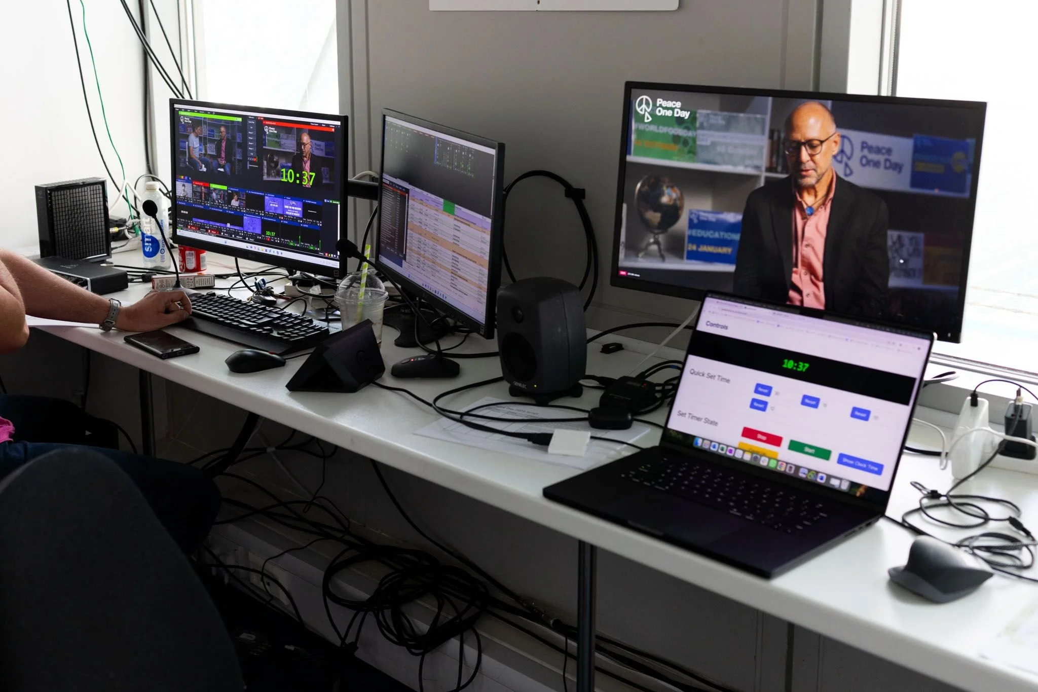 Multiple monitors on a white desk showing a video interview, editing software, and a clock reading 10:37. There is a calendar on a laptop screen, a keyboard, mouse, and various electronic devices on the desk. Behind the monitors, there is white wall and window, with cables and a small speaker.