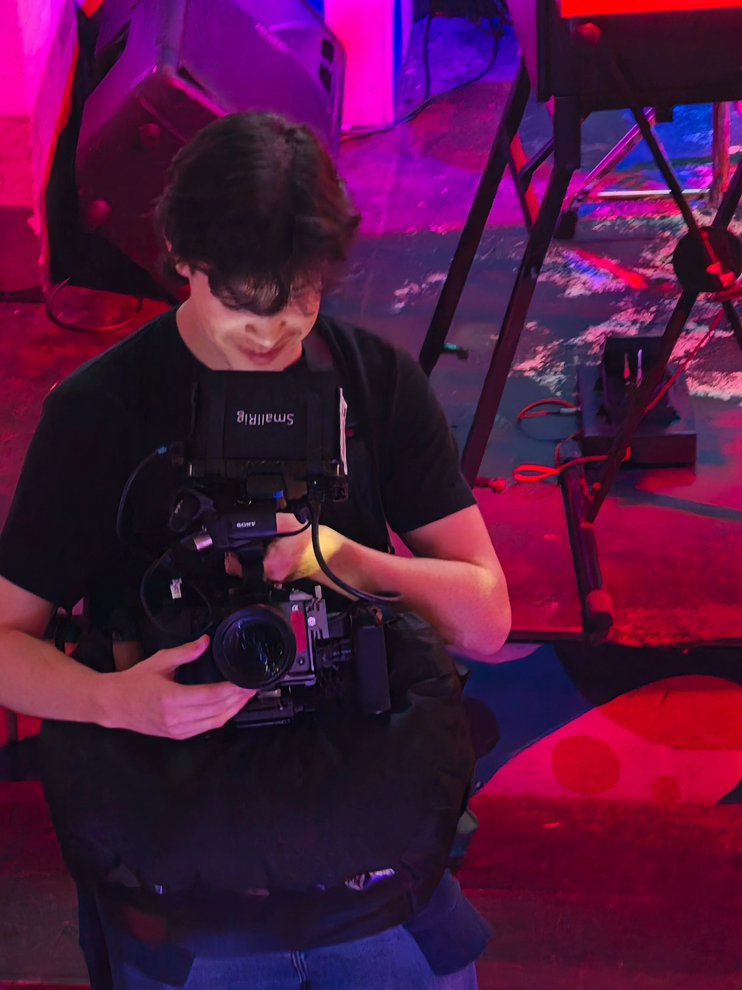 A person with medium-length dark hair taking a photo using a vintage camera, in a setting with colorful red and purple lighting.