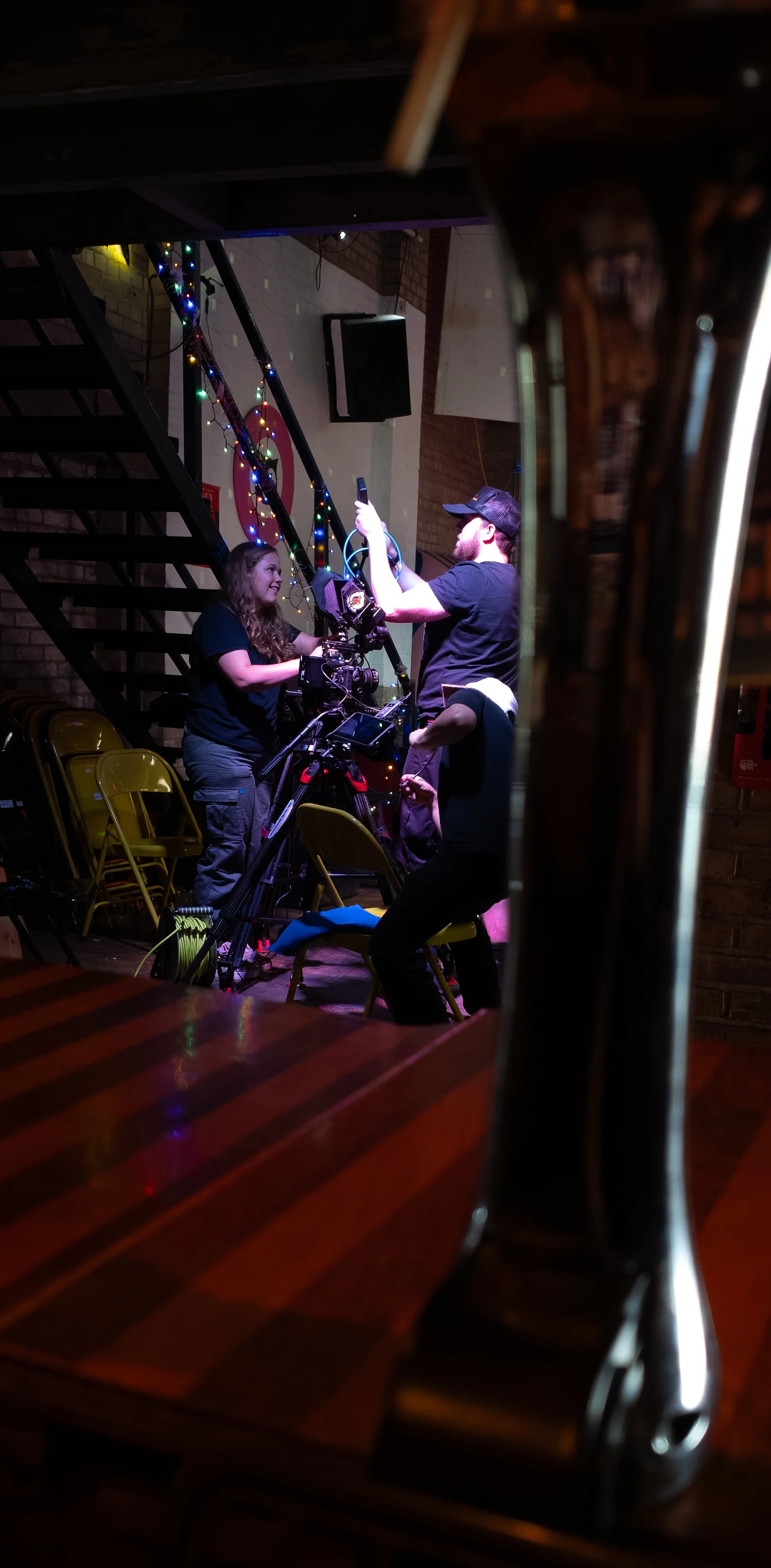 Two people behind the scenes at a film or video shoot, with equipment and cameras, seen through a mirror or reflective surface in a dimly lit venue. One person is smiling, and both are focused on the equipment and filming process. Decorations like string lights are in the background.