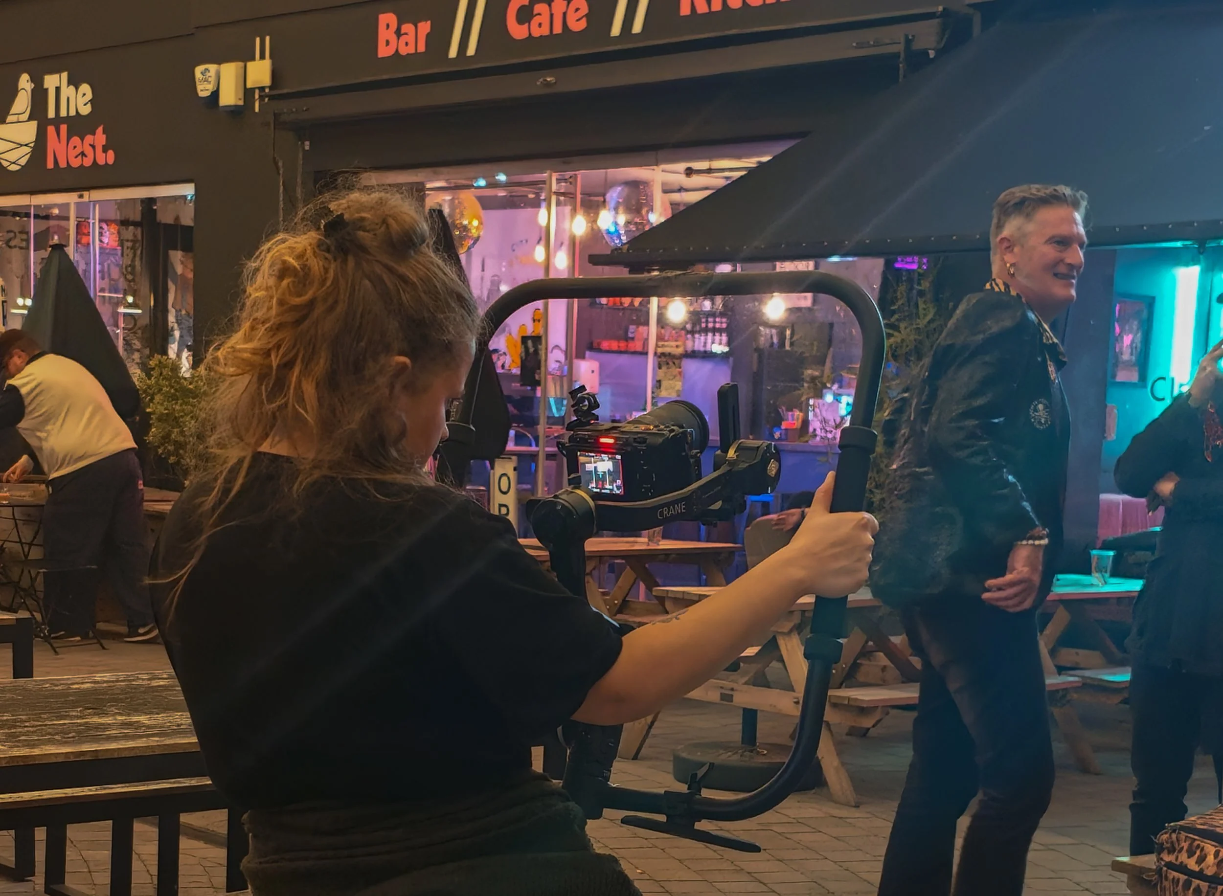 A woman with curly hair operating a camera on a stabilizer at an outdoor venue during nighttime. Behind her, a smiling person with short hair and earrings is engaging with others. The background shows a storefront with illuminated signs and decorations, suggesting a lively, social atmosphere.