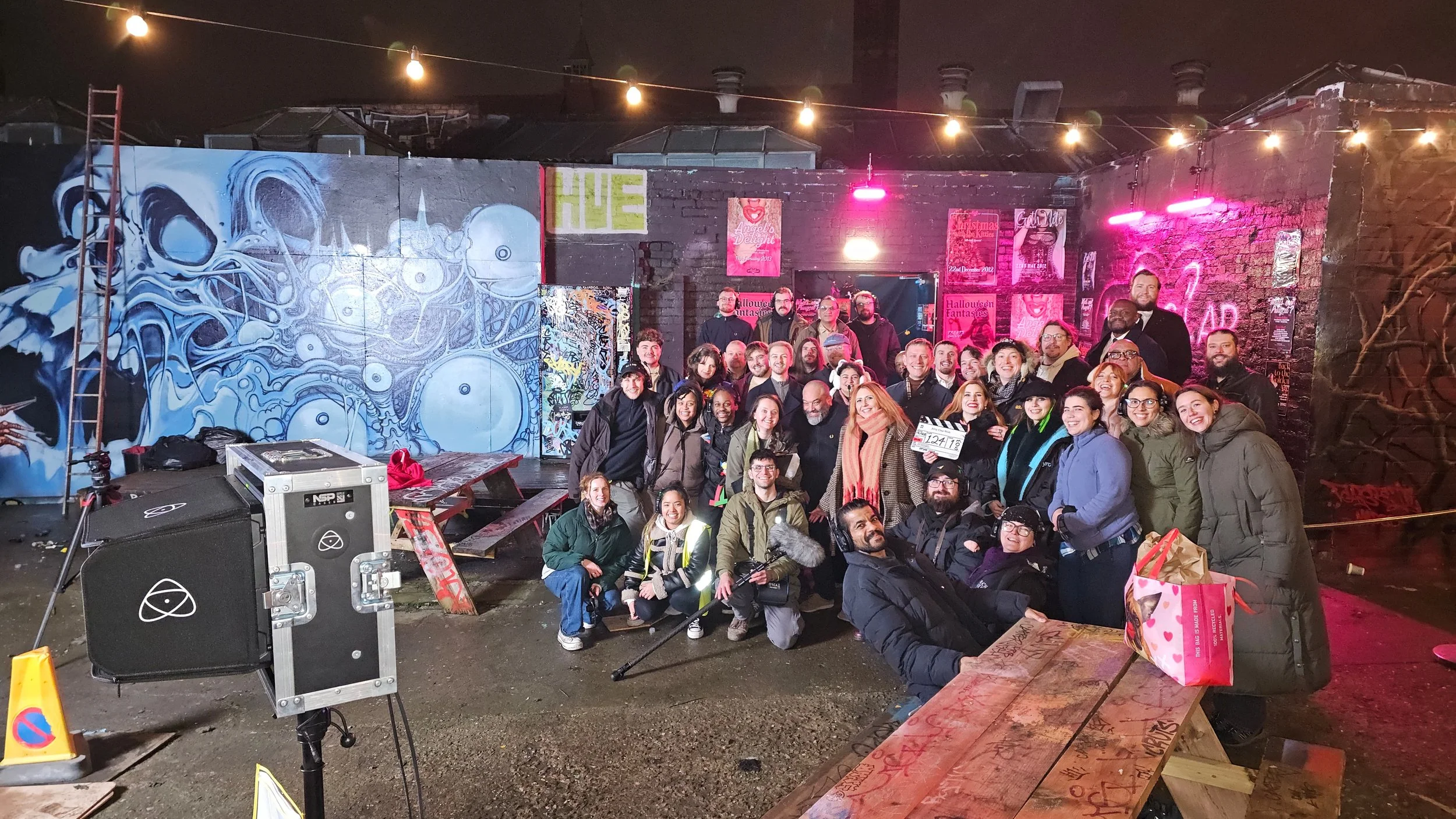 Group of people gathered in an outdoor space at night, with graffiti art on the brick wall behind them, graffiti posters, and string lights overhead, smiling and posing for a photo.