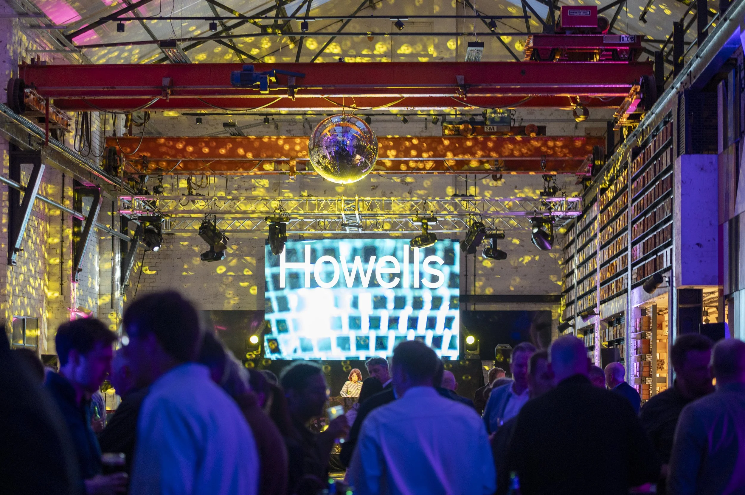 A crowd of people at a lively event with a disco ball hanging from the ceiling, colorful lights, and the word "Howells" displayed on a screen amidst technical equipment.