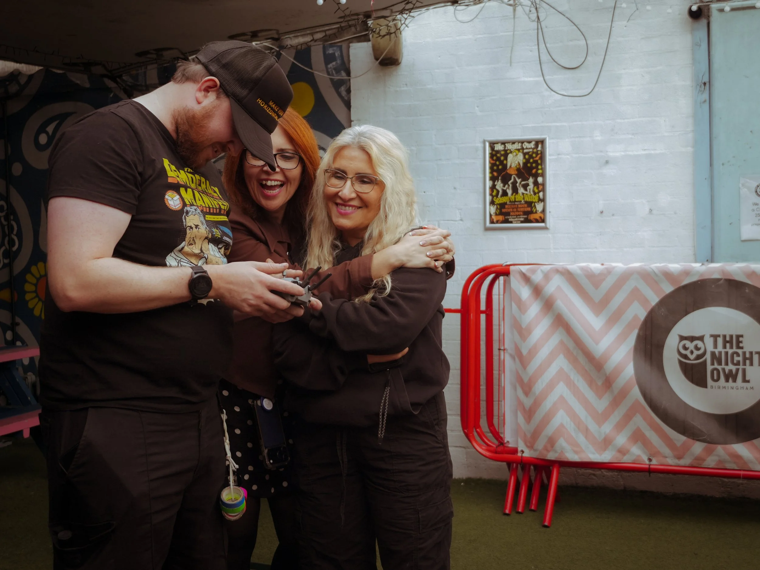 Three people, two women and a man, are smiling and looking at a remote control, standing close together in a room with a white brick wall decorated with posters. One woman has blonde hair and glasses, the other has red hair and glasses, and the man has a beard and is wearing a baseball cap and a t-shirt with a character on it.