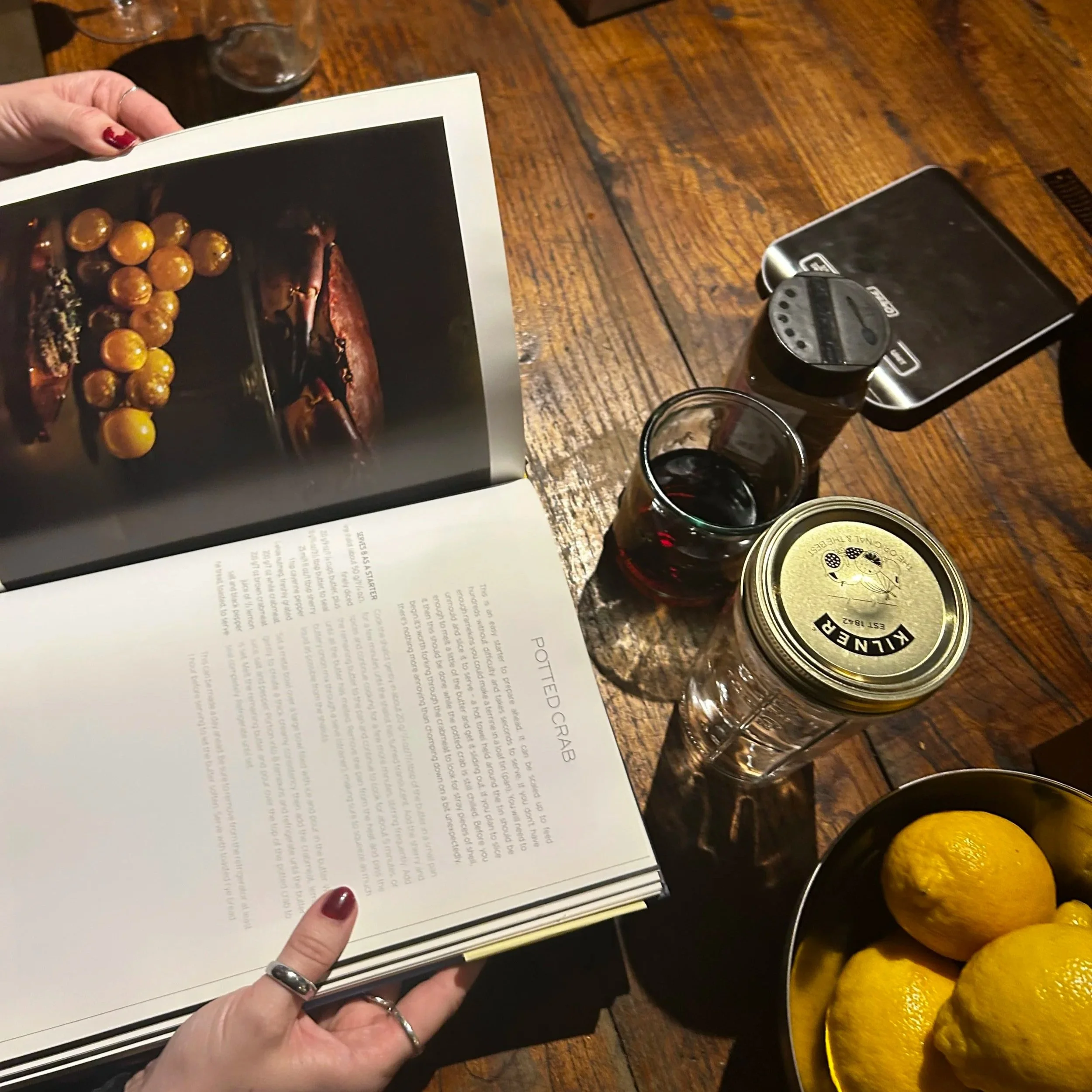 A person with red painted nails holding an open book over a wooden table, with a glass of red wine, a plate of lemons, and an electronic device with a camera on the table.