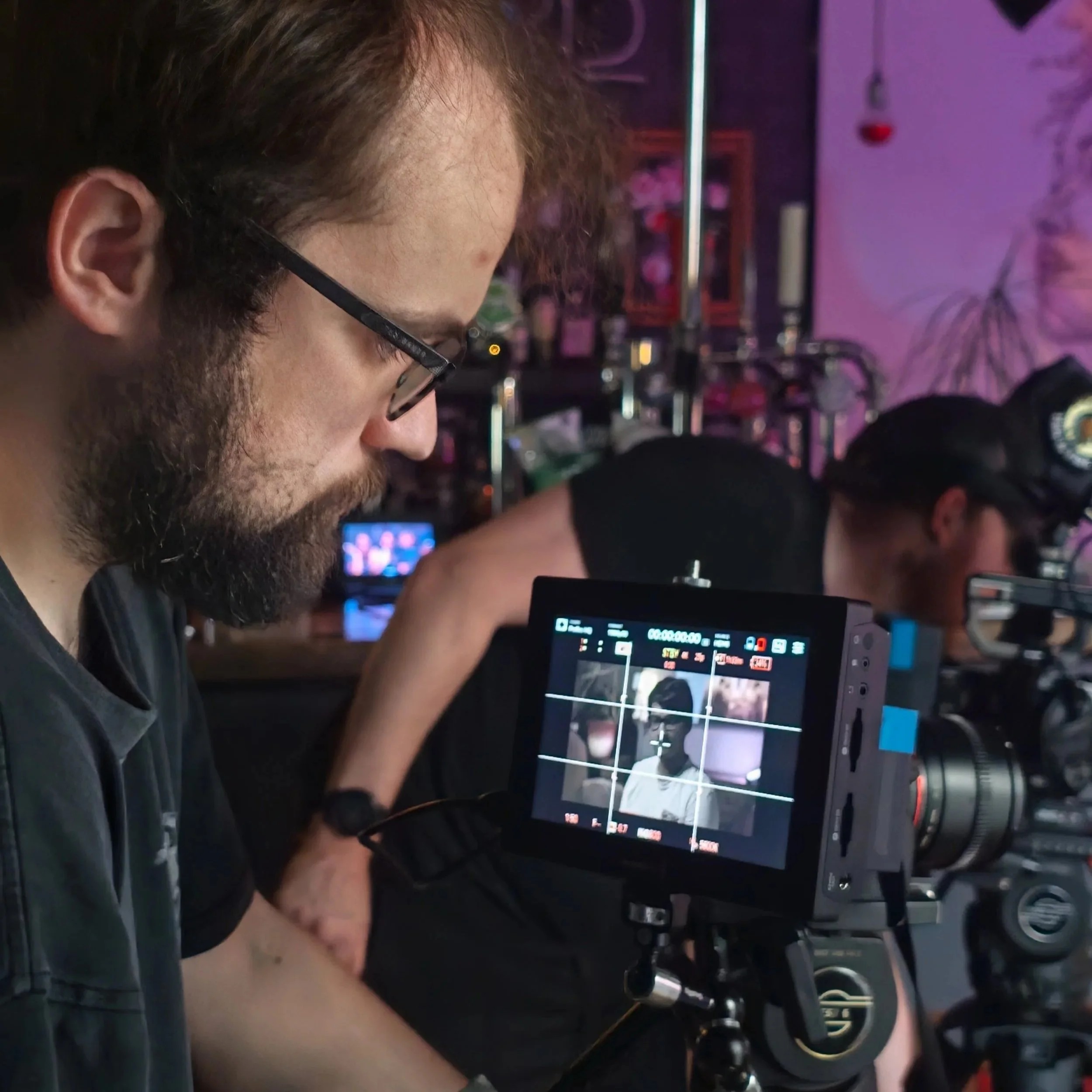 A man with glasses and a beard looking down at a camera viewfinder, with a background of neon purple lighting and shelves filled with various objects.