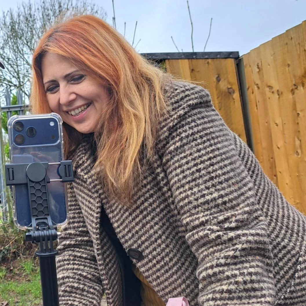 A woman with long red hair is smiling and filming something on a phone that is mounted on a tripod. She is wearing a brown and black checkered coat, and there is a wooden fence and some trees in the background.