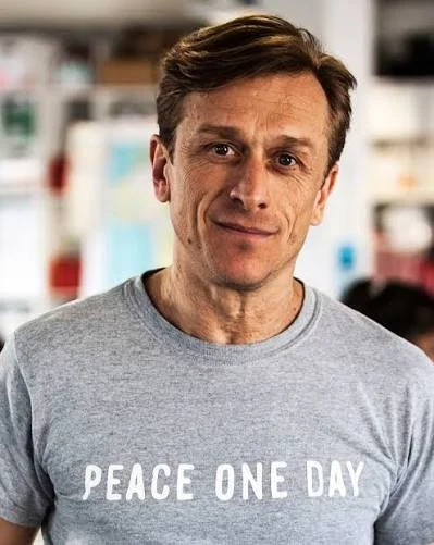 A man wearing a gray t-shirt with the words "PEACE ONE DAY" printed on it, standing indoors with a blurred background.