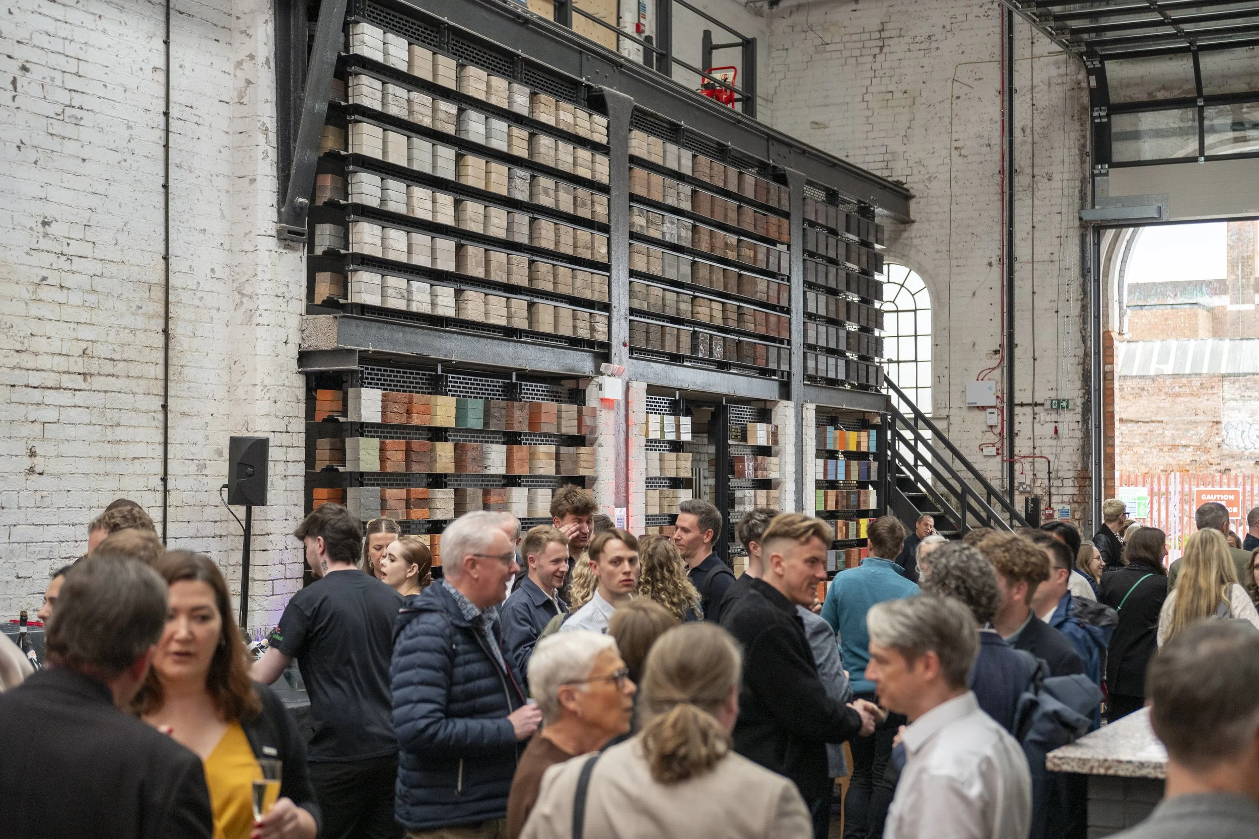 Crowd of people gathered inside a spacious industrial-style venue with white brick walls and large windows, with a display of various color and pattern samples on a tiered metal shelving unit.