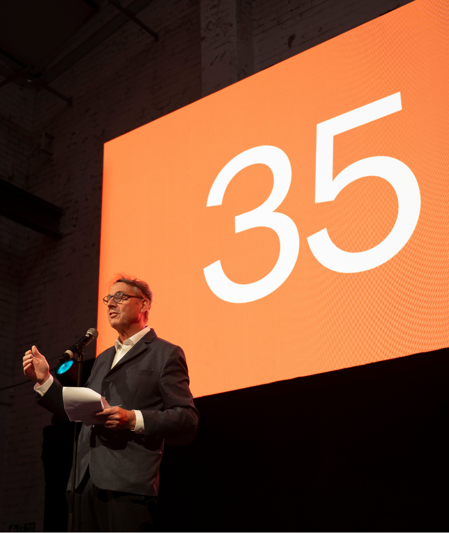 A man in a suit speaking into a microphone on stage with a large orange screen displaying the number 35.