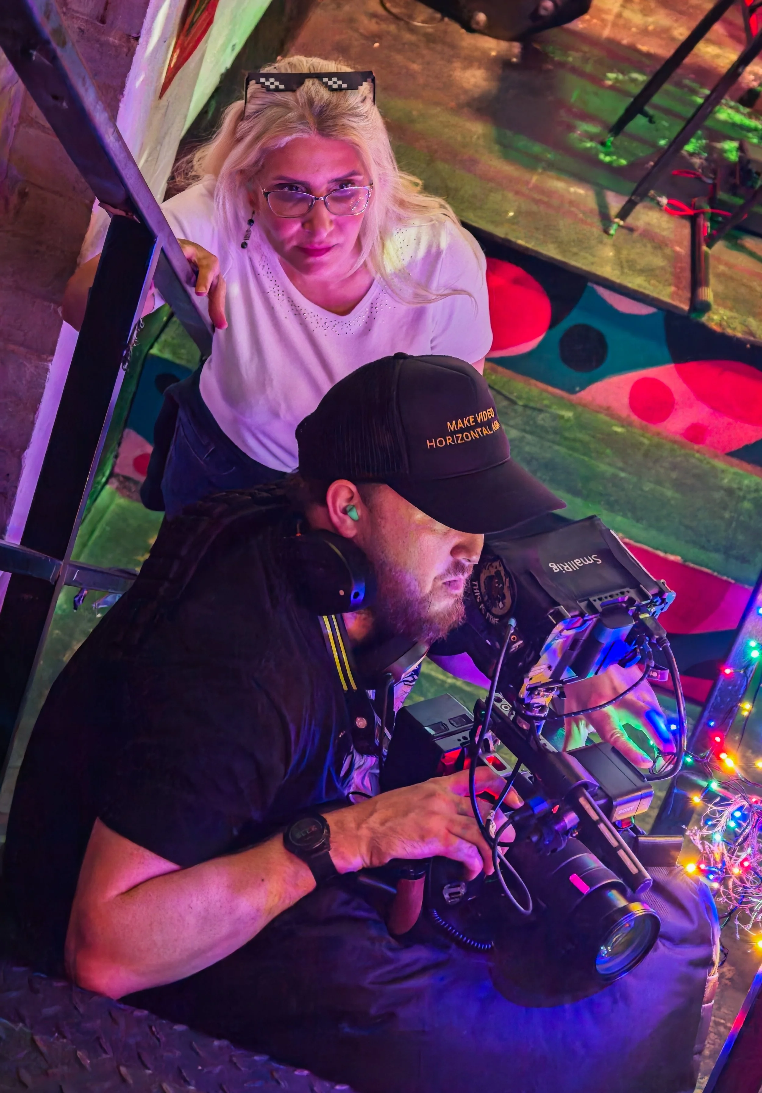 A woman with blonde hair, glasses, and a white shirt leans over a DJ mixing equipment decorated with colorful string lights, while a man in a black cap and headphones works on the gear in a vibrant, colorful setting.