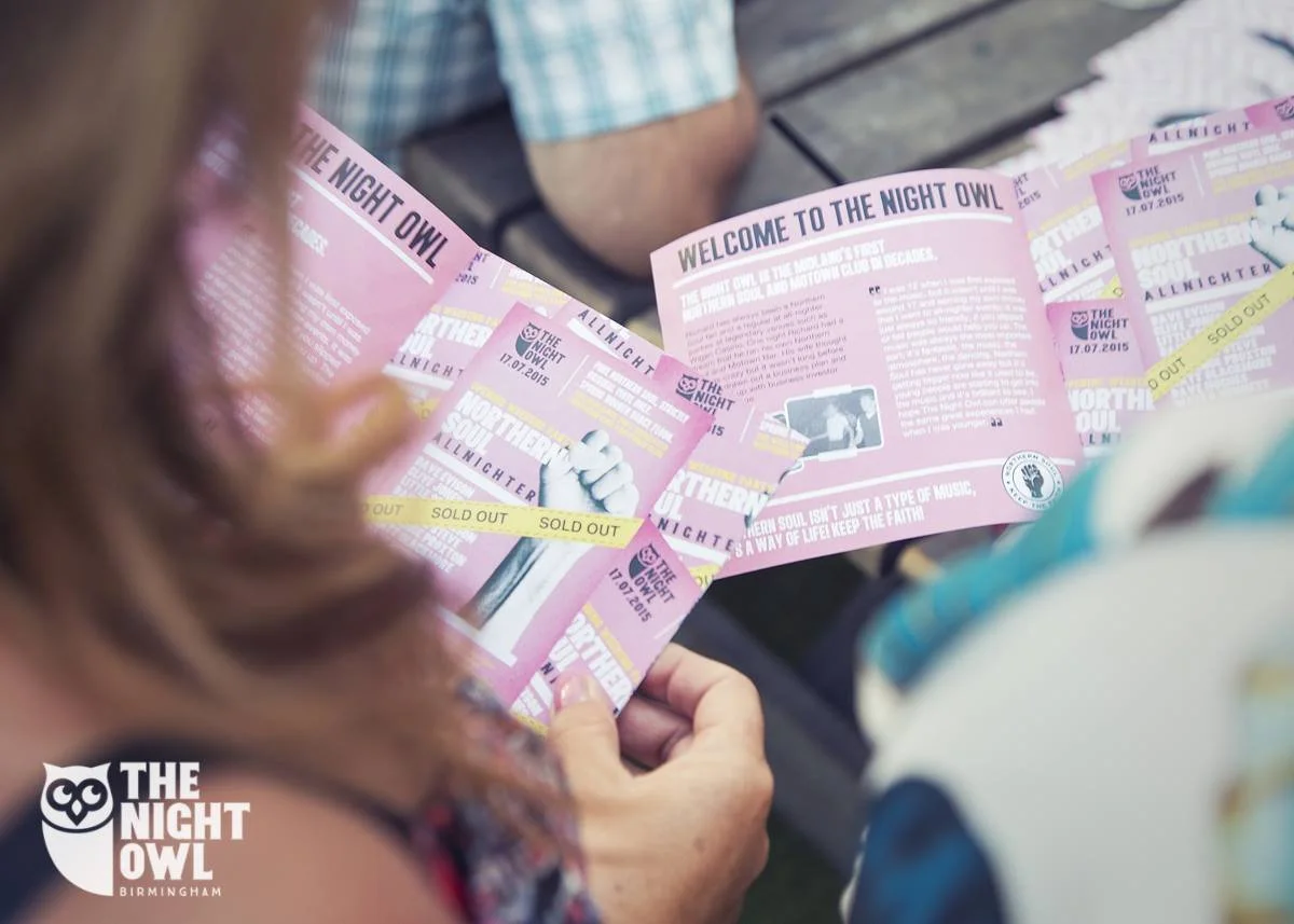 Person holding pink event flyers for 'The Night Owl' music event, with 'Sold Out' tape across some flyers, at an outdoor gathering.