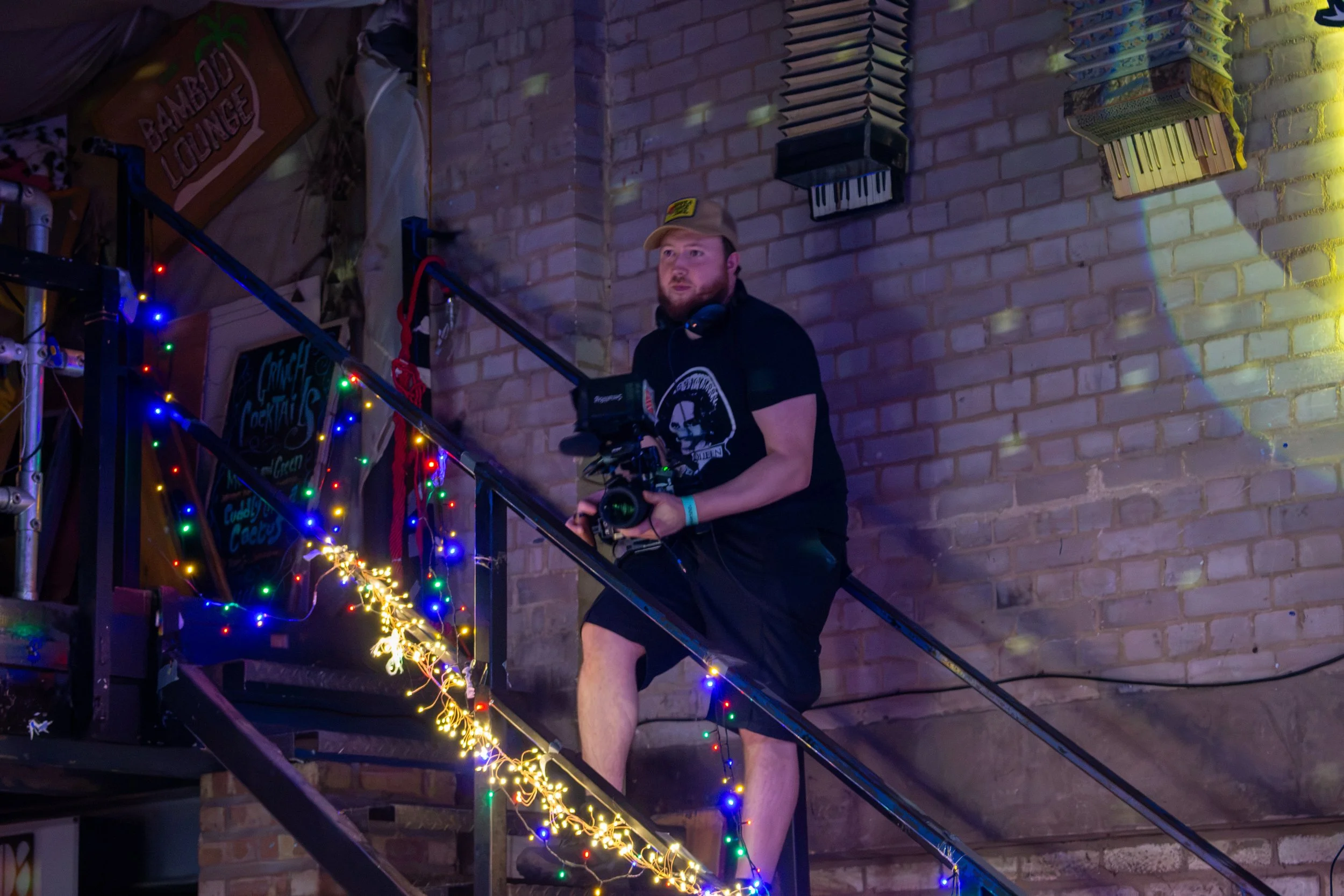 A man with a beard and wearing a cap and headphones around his neck is standing on a staircase decorated with colorful string lights. He is holding a camera and looking slightly to his right. The background features a brick wall with music-themed wall decorations including a piano and a sign that reads 'Bamboo Lounge'.