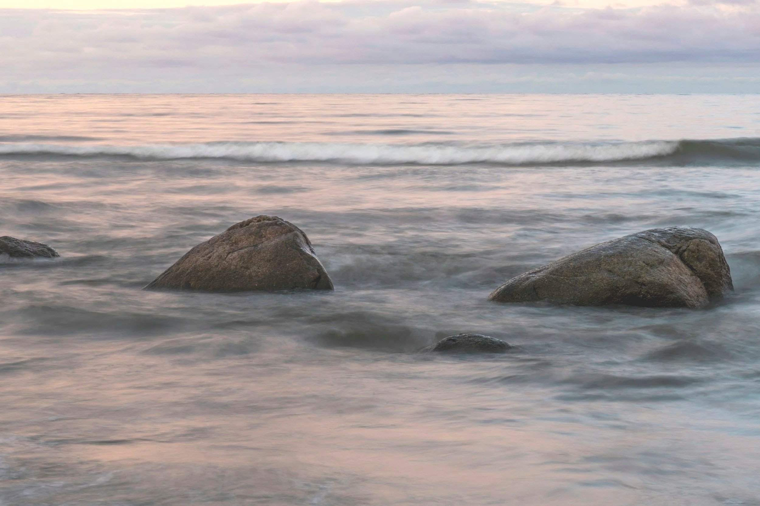 Seaside scene with three large rocks, representing parts of an ecosystem, in shallow water, gentle waves, and a cloudy sky at dusk.