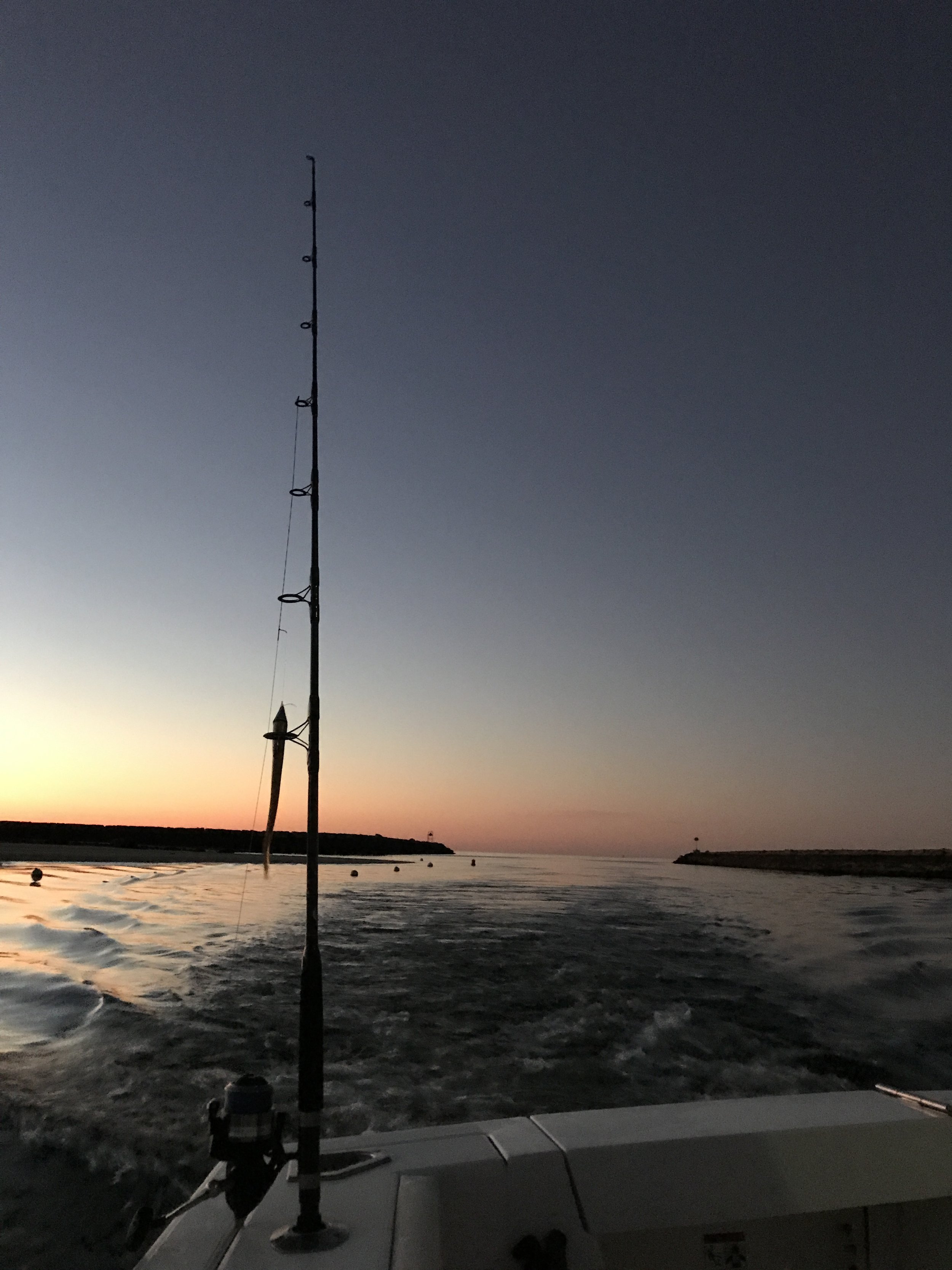 Fishing rod with a fishing reel on a boat at sunset by the ocean, with a shoreline and small lighthouse in the distance.
