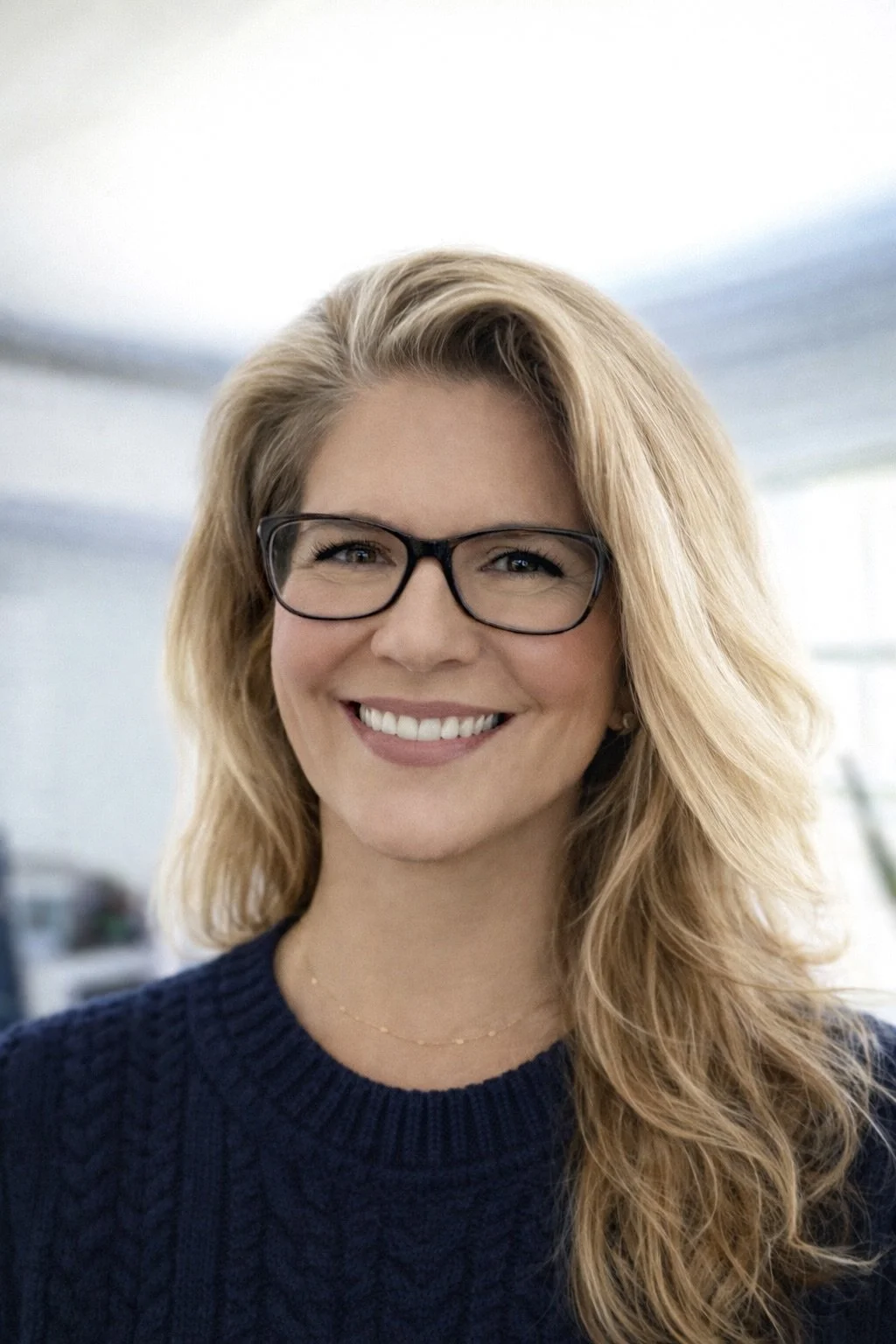 A woman with blonde hair, glasses, and a navy sweater smiling in an indoor setting.