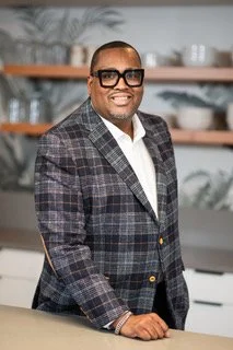 A man in a black suit and white shirt sitting and smiling in an indoor setting