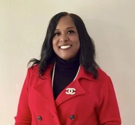 A woman with shoulder-length black hair smiling, wearing a black blazer, sitting on a beige chair in a neutral-toned room.