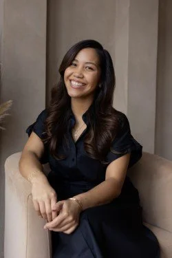 A woman with long dark hair, wearing a black top, sitting on a beige sofa and smiling at the camera.