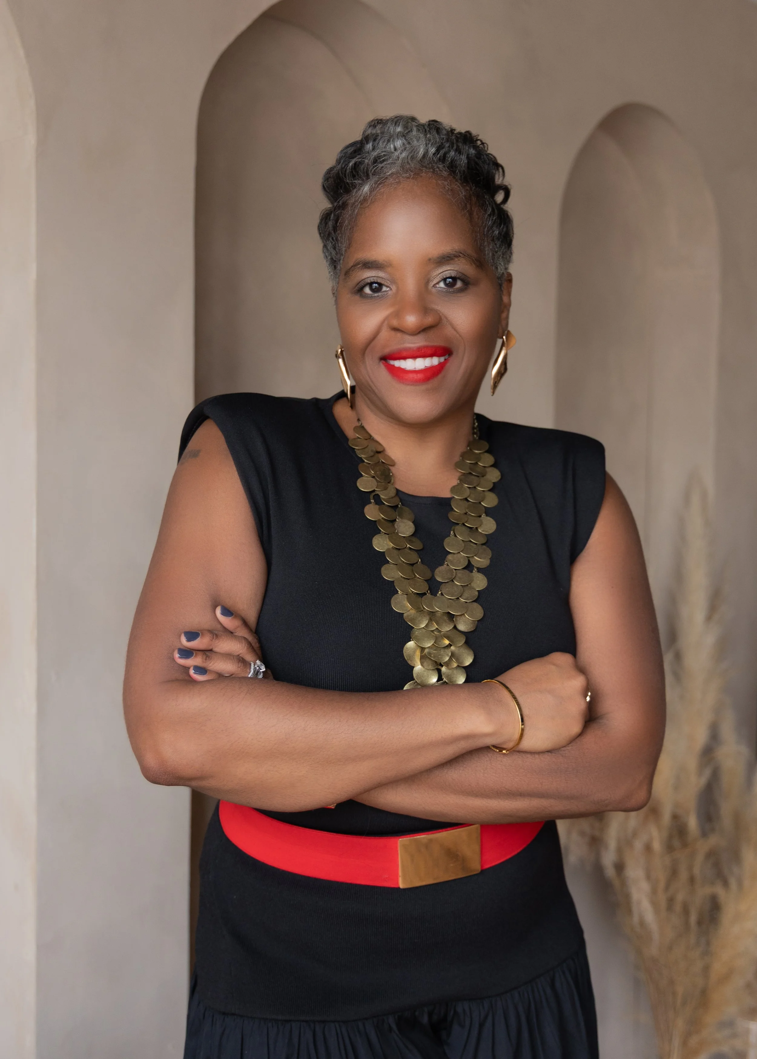 Smiling woman with short curly hair wearing a black tank top and necklace, standing outdoors with blurred greenery and blue flowers in the background.