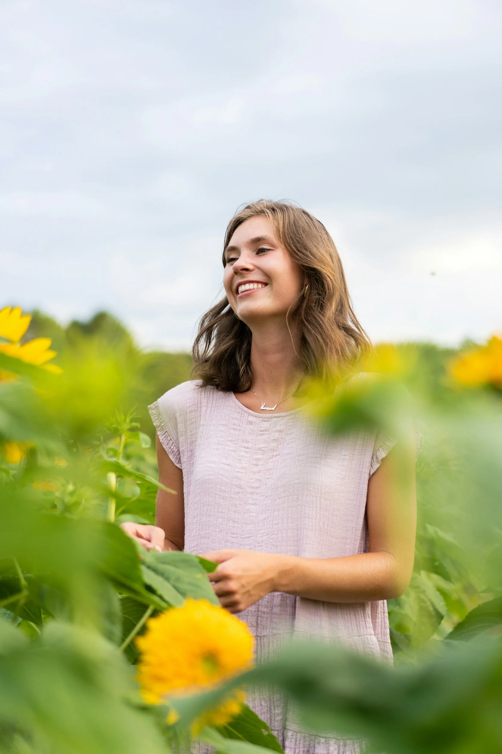 A young woman smiling outdoors in a field of yellow flowers under a cloudy sky.