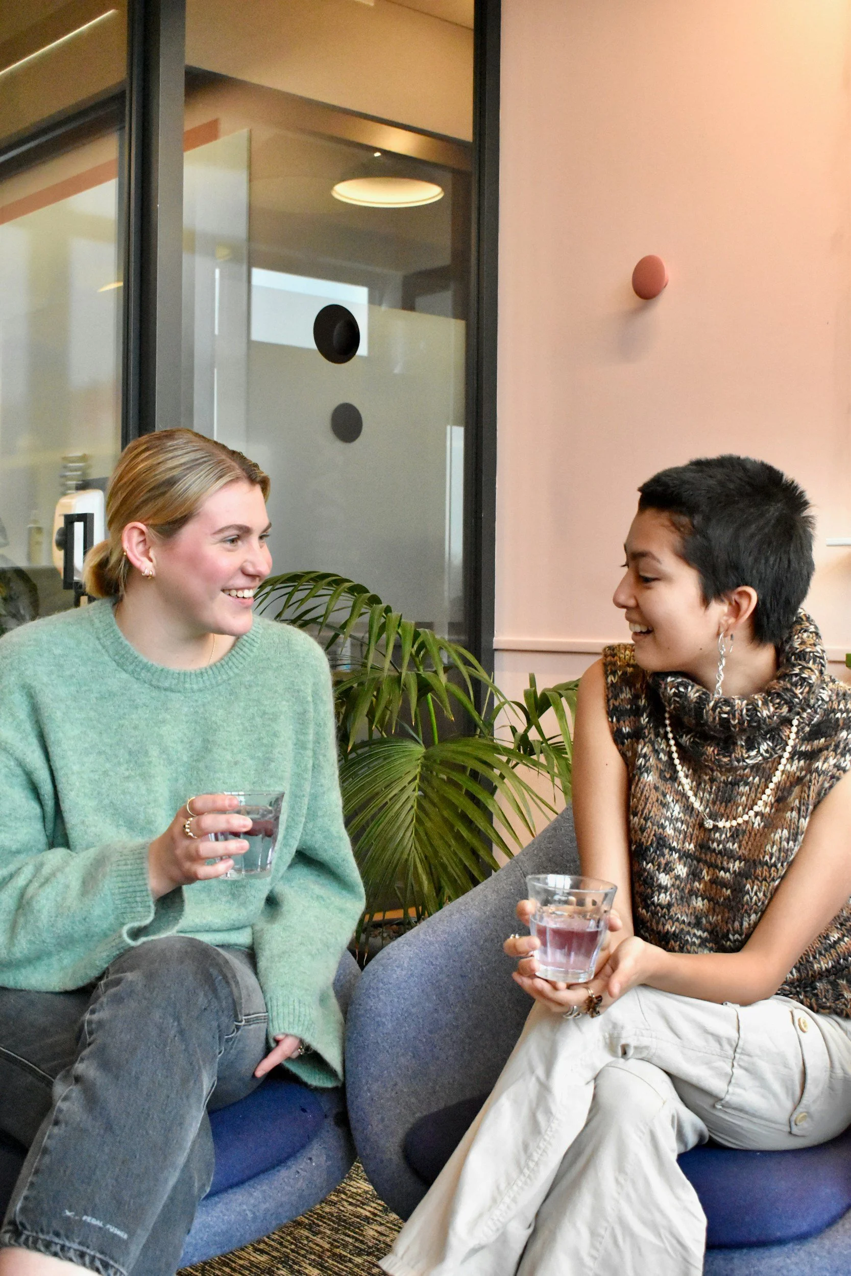 Two women sitting on round chairs, holding glasses of water, and engaging in a conversation in a modern indoor setting with a glass wall and green plants.