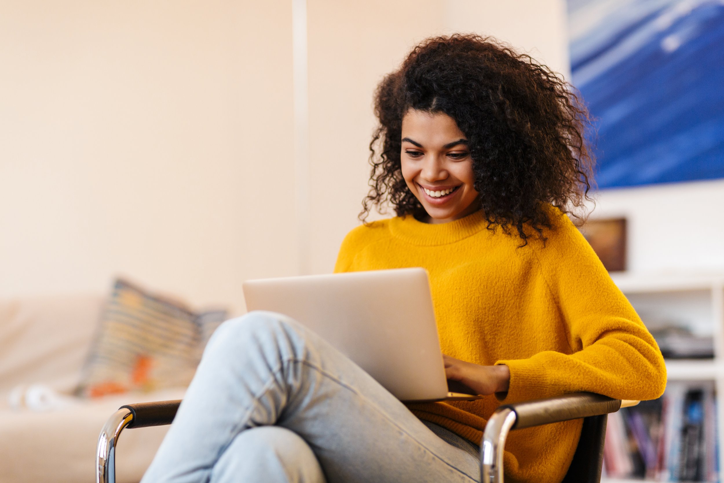 A young woman with curly hair wearing a yellow sweater, sitting in a chair, looking at her laptop and smiling.