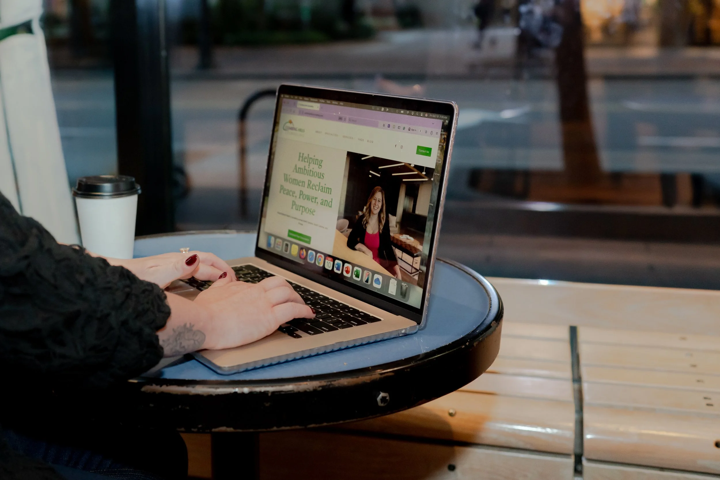 Person working on a laptop at an outdoor café table with a disposable coffee cup, looking at a website promoting empowering women.