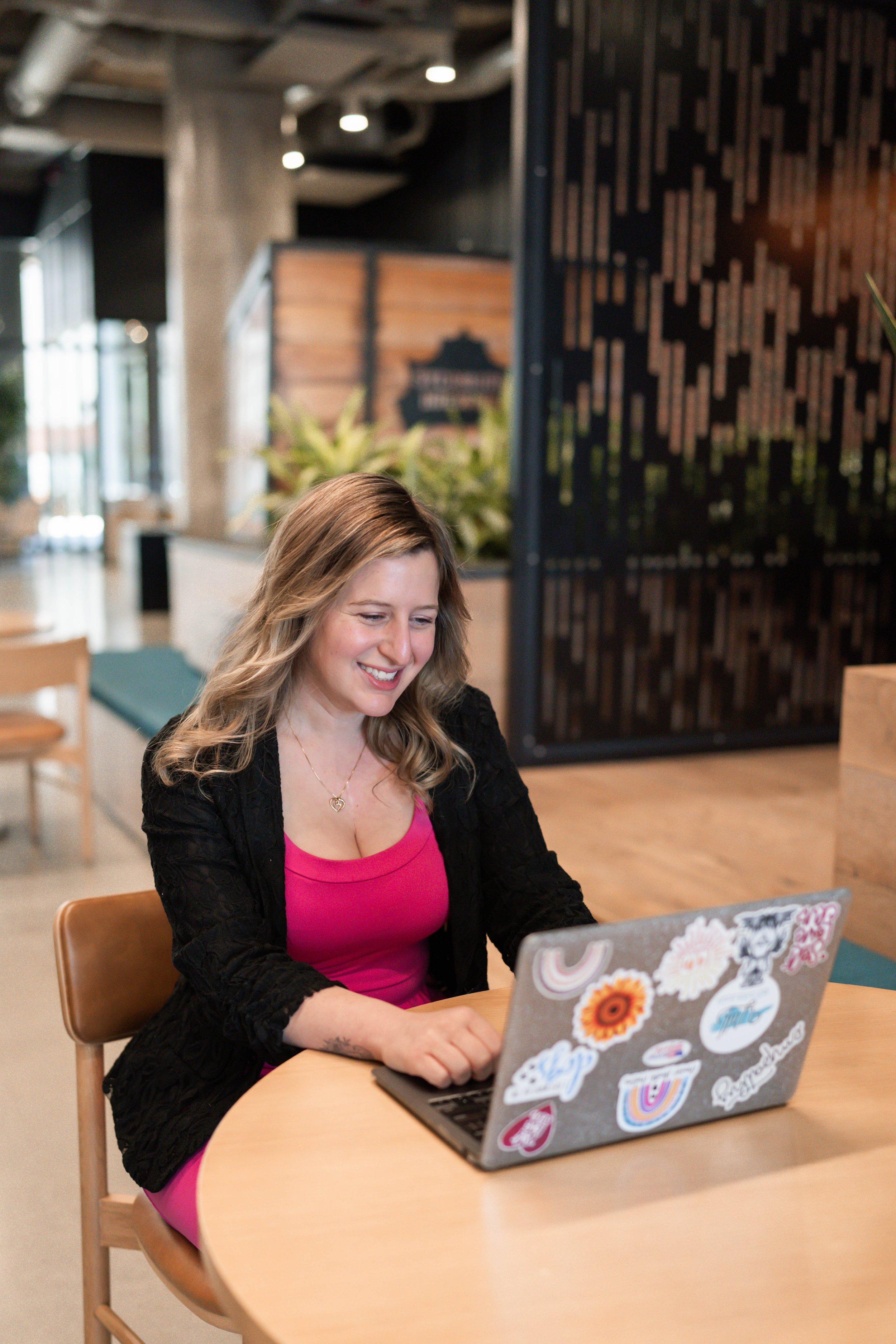 A woman sitting at a round wooden table, smiling while looking at her laptop, decorated with various stickers, in a modern indoor setting with plants and wooden accents.