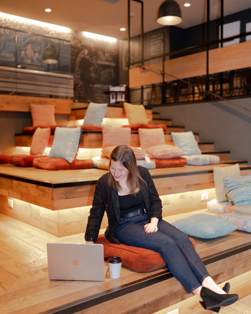 A woman sitting on a cushion on a tiered wooden seating area, smiling and looking at a laptop, with a coffee cup next to her, in a modern cafe or lounge with colorful pillows and chalkboard walls.