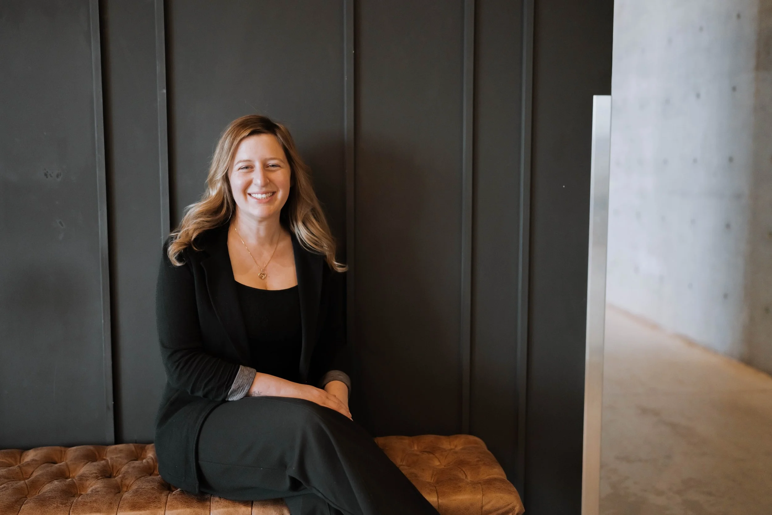 A woman with long wavy brown hair, wearing a black blazer and black pants, sitting on a tufted brown leather bench, smiling against a dark wall in an indoor setting.