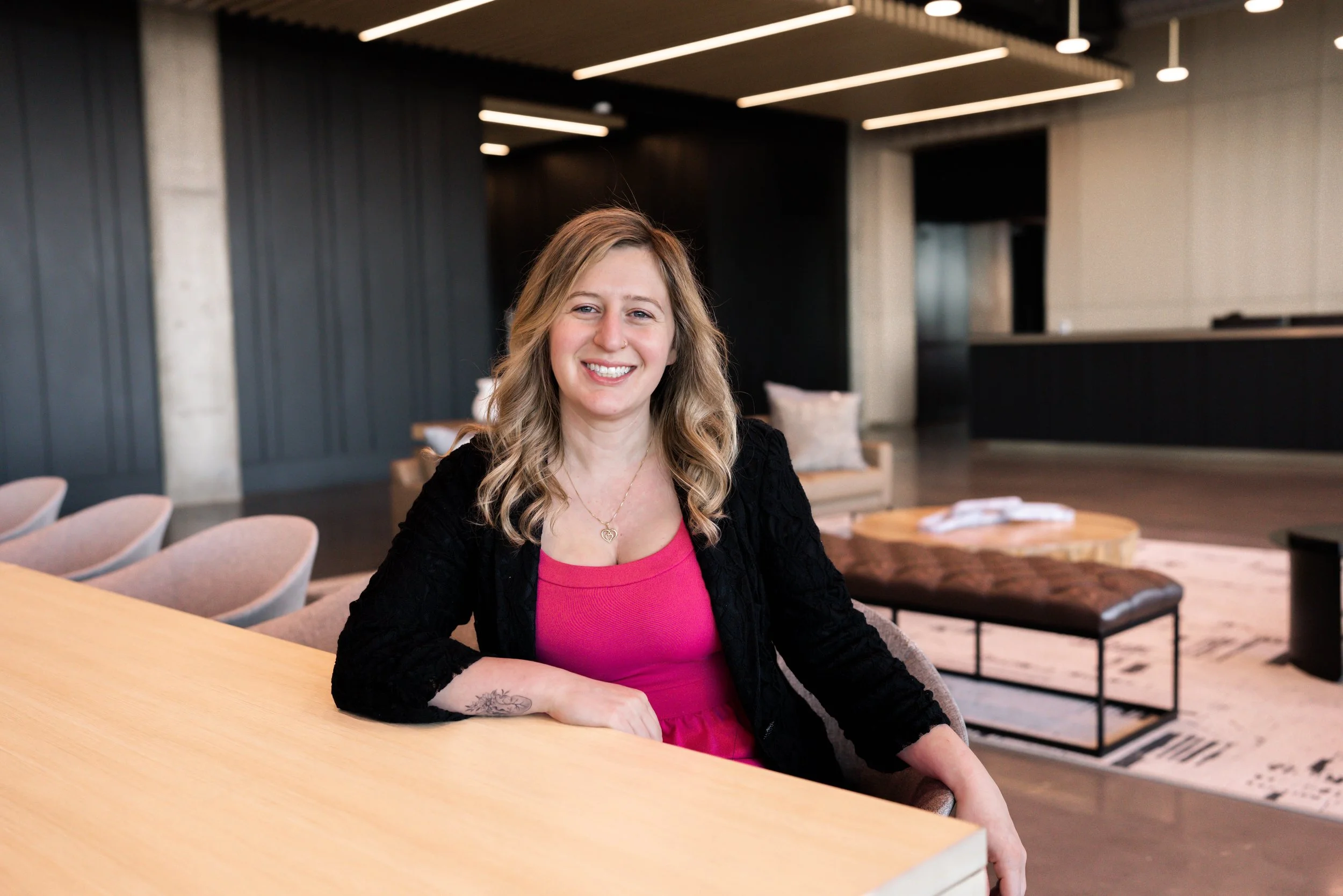 A woman with blonde hair, smiling, seated in a modern lounge or hotel lobby, wearing a pink shirt and black blazer, with a tattoo on her right wrist, in a space with contemporary furniture and dark wall paneling.