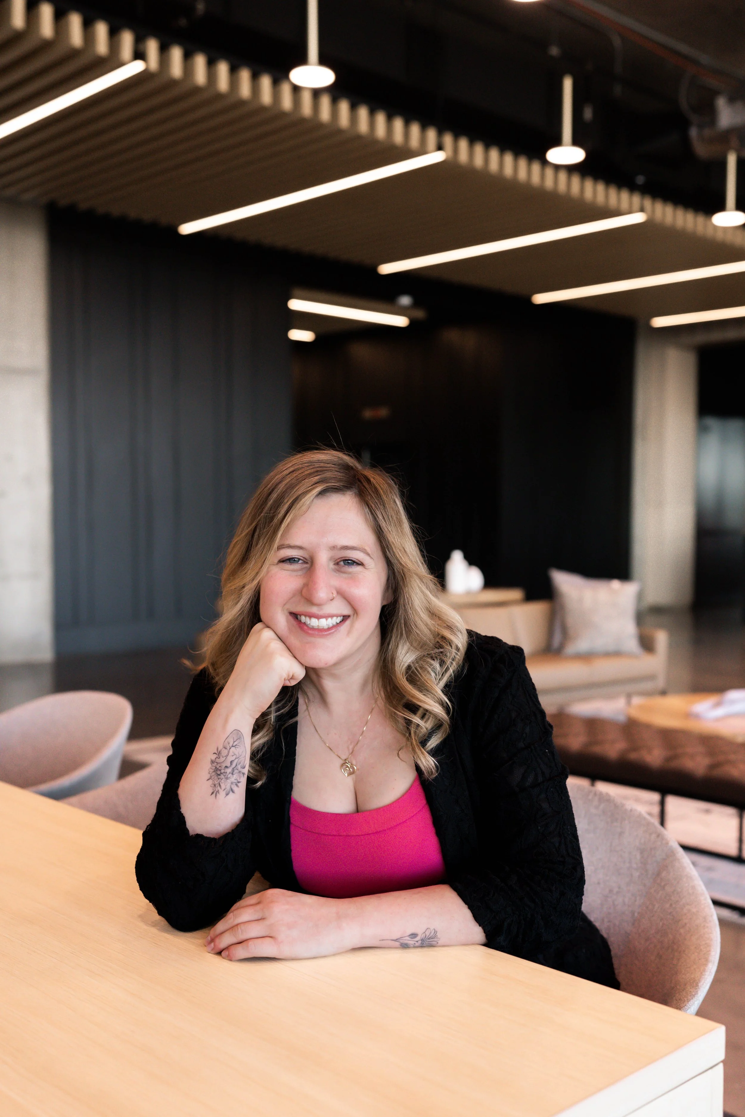 A woman with blonde hair, wearing a pink top and black cardigan, smiling at the camera while sitting at a wooden table in a modern, well-lit room with black and beige decor.