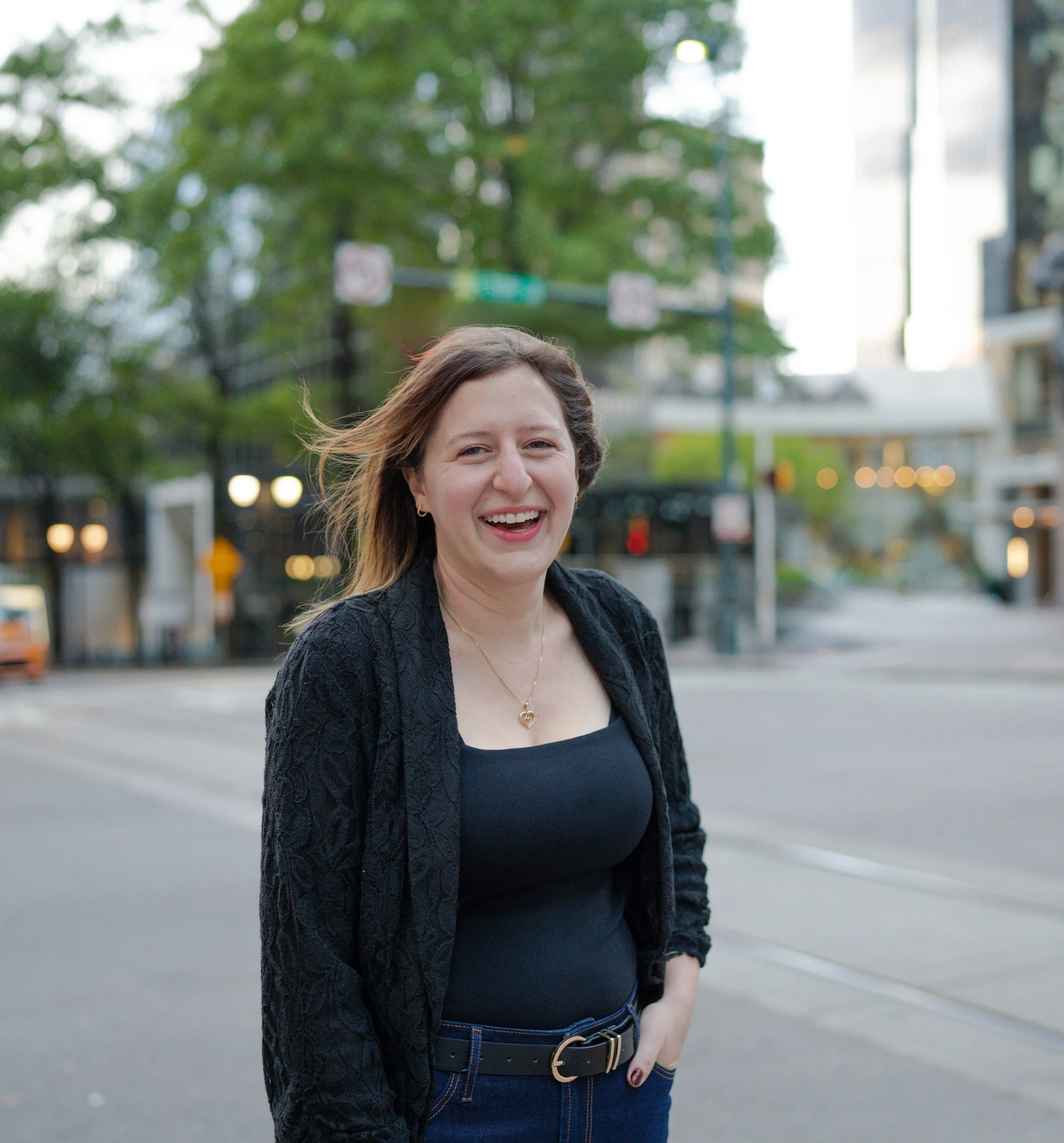 A woman with shoulder-length light brown hair smiling on a city street, wearing a black top, patterned black jacket, and jeans, with trees and buildings in the background.